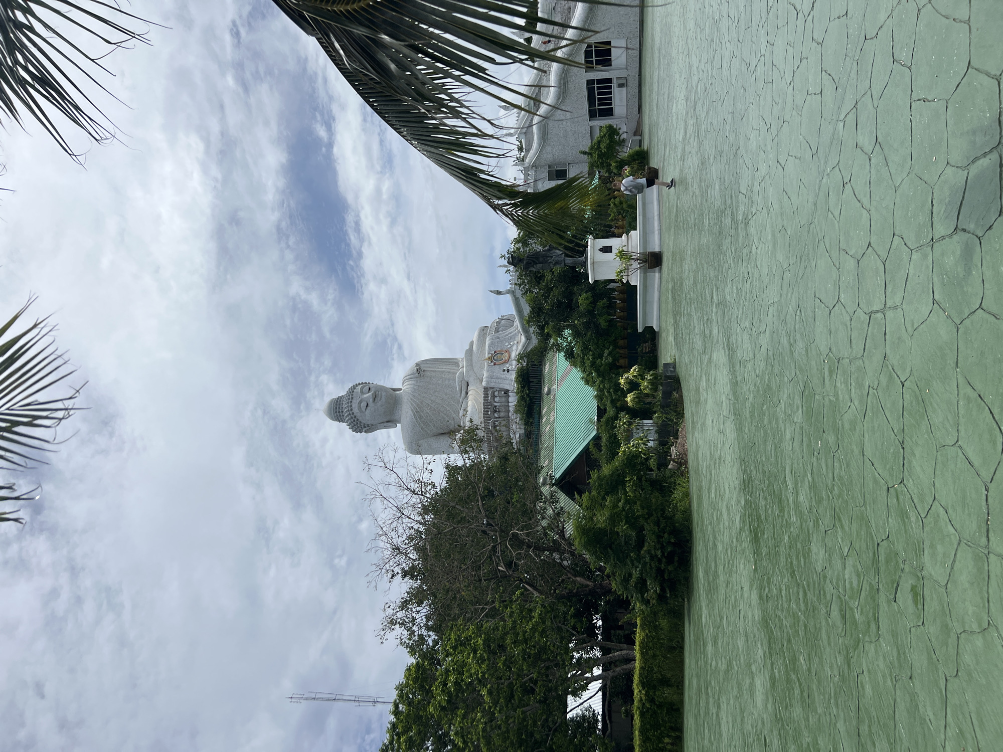 In Rawai, Thailand, a large, serene white Buddha statue is perched atop a hill. The statue faces forward with its eyes closed in meditation. In the foreground, a person walks across a green, textured courtyard. The courtyard features a patterned floor made of irregular polygonal shapes. Behind the person, a bronze statue stands on a white pedestal. Lush green trees and buildings with green roofs surround the base of the Buddha statue. Palm fronds frame the top of the image, and a cloudy sky stretches above. A metal tower stands to the left of the Buddha statue.
