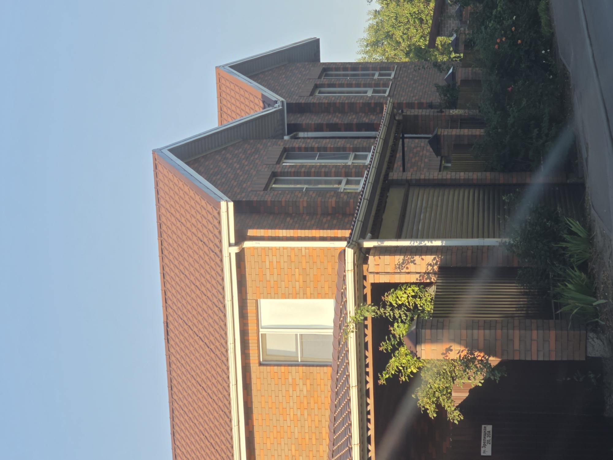 The image shows a two-story house constructed with brick in a suburban setting. The house has a brown tiled roof, multiple windows with white frames, and a covered porch area. Adjacent to the house is a fence made of brick pillars and vertical metal slats. A dark brown gate with a sign that reads "ул. Зрелищная 20, 20А" is visible to the left. Green vegetation, including trees and bushes, surrounds the property. The scene is illuminated by natural light, suggesting it was taken during the day. The sky is clear and blue. The overall impression is of a residential property.