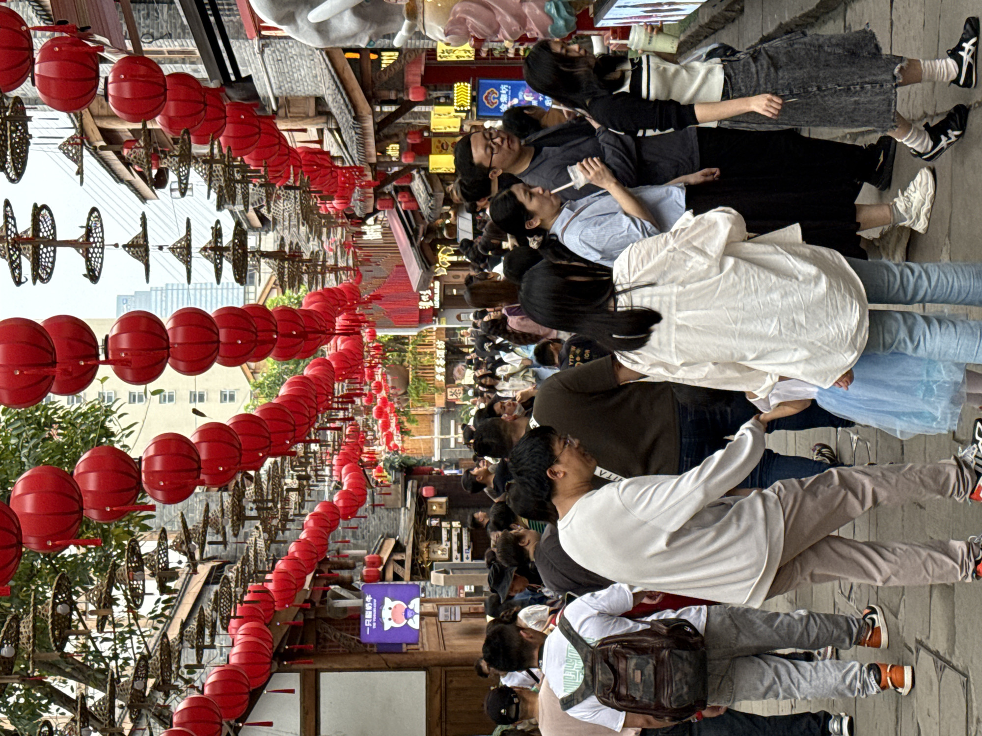 The image shows a crowded street scene in Yuzhong District, China. Red lanterns and ornate bronze fixtures hang above the street. A diverse group of people walks along the cobblestone pavement. Many are in casual clothing. A building is on both sides of the street. A modern high-rise is visible in the distance.
