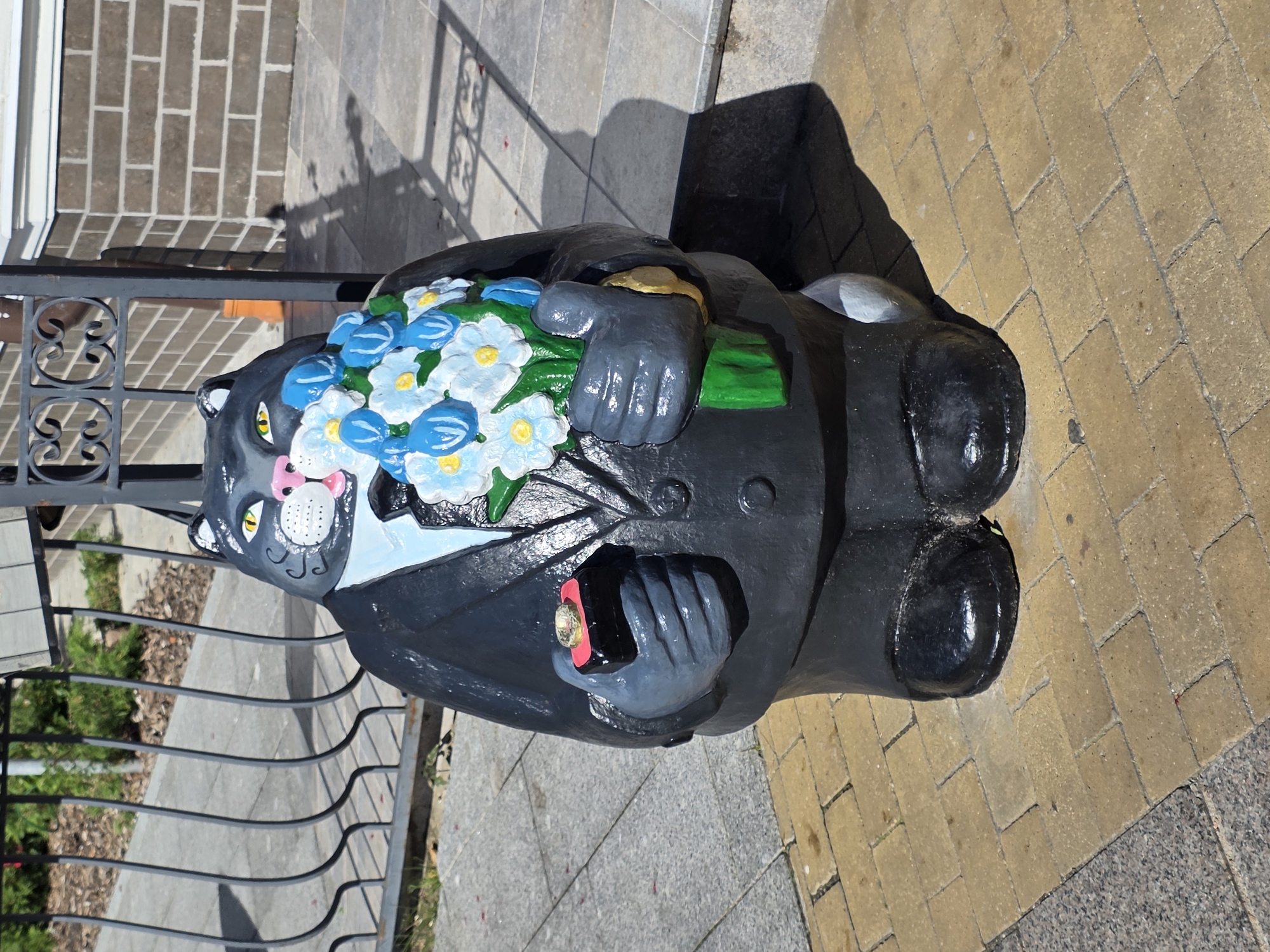 A concrete sculpture of a gray cat wearing a black suit, white shirt, and black tie stands on a paved area in front of a building. The cat holds a bouquet of blue and white flowers in its right paw and a small red box containing a ring in its left paw. The building behind the sculpture is made of brick and has a decorative metal fence. The ground is paved with yellow bricks. The scene appears to be in Zelenogradsk, Russia.