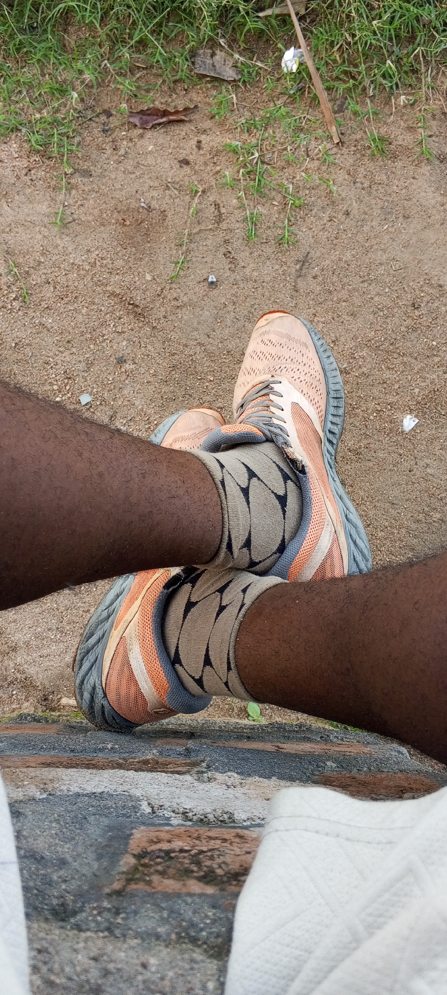 The image shows a close-up, overhead view of a person's legs and feet, positioned on a low, rough-textured stone wall. The person is wearing athletic shoes, primarily light orange with grey and dark grey accents, and patterned socks that are a light tan with dark triangular shapes. Their legs are dark-skinned and appear to have light stubble. The background consists of a sandy area with sparse green grass and a few scattered dry leaves and debris. The scene is outdoors, on what appears to be a dirt or gravel path next to a grassy area. The lighting suggests it is daytime. The image does not contain any identifiable landmarks or specific elements that would allow for precise localization within Jalingo, Nigeria, beyond the general context of an outdoor, possibly recreational or transitional space.