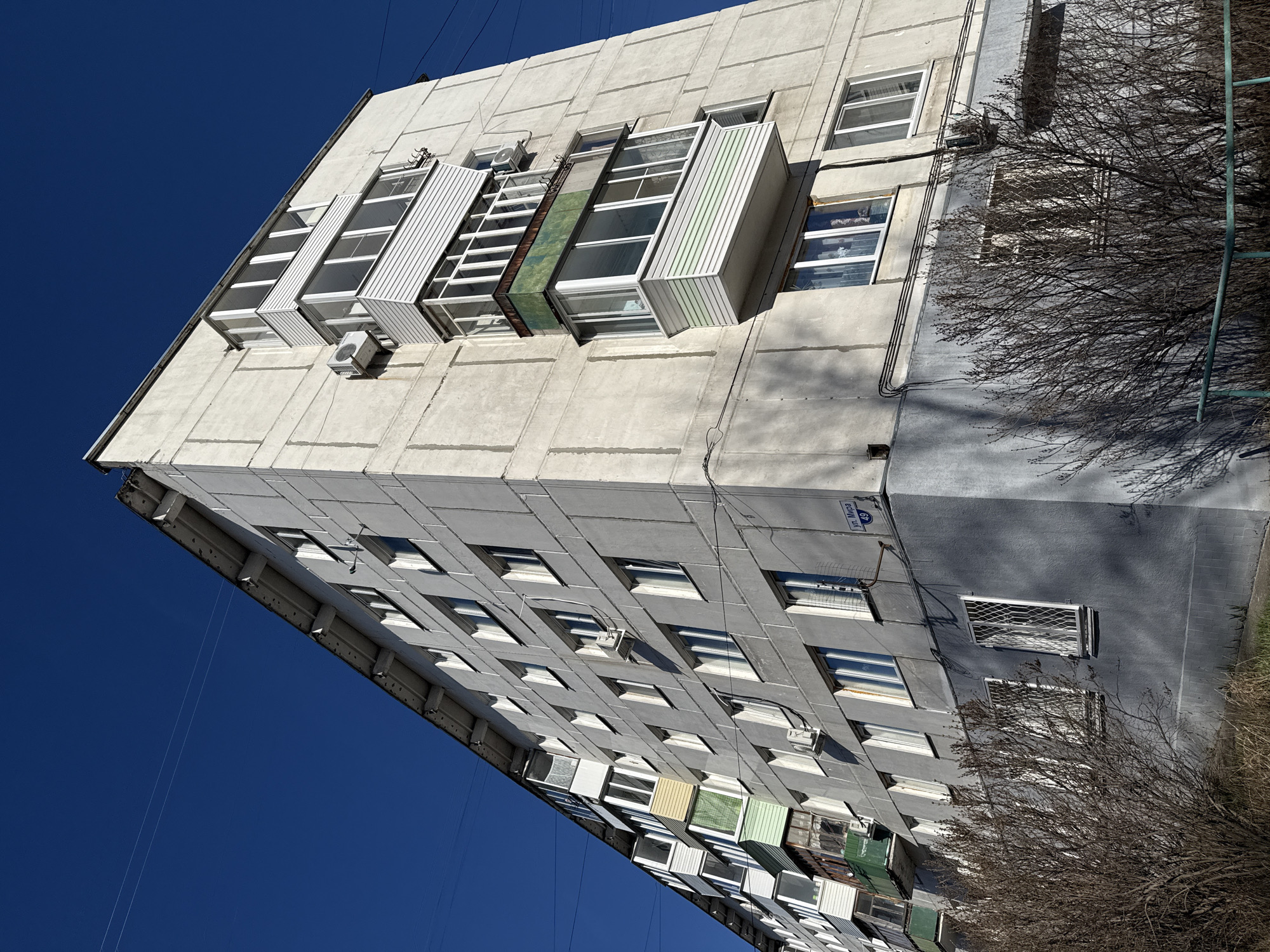 A low-angle, medium shot depicts a tall, gray, concrete apartment building against a bright blue sky. The building's facade is characterized by a grid of rectangular windows, many with air conditioning units installed beneath them. Some windows are flanked by protruding, box-like structures, resembling small, enclosed balconies. On the right side of the building, bare tree branches partially obscure the view. A small sign on the building reads "Ул. Мира, 49". Wires are visible extending from the top of the building into the sky.