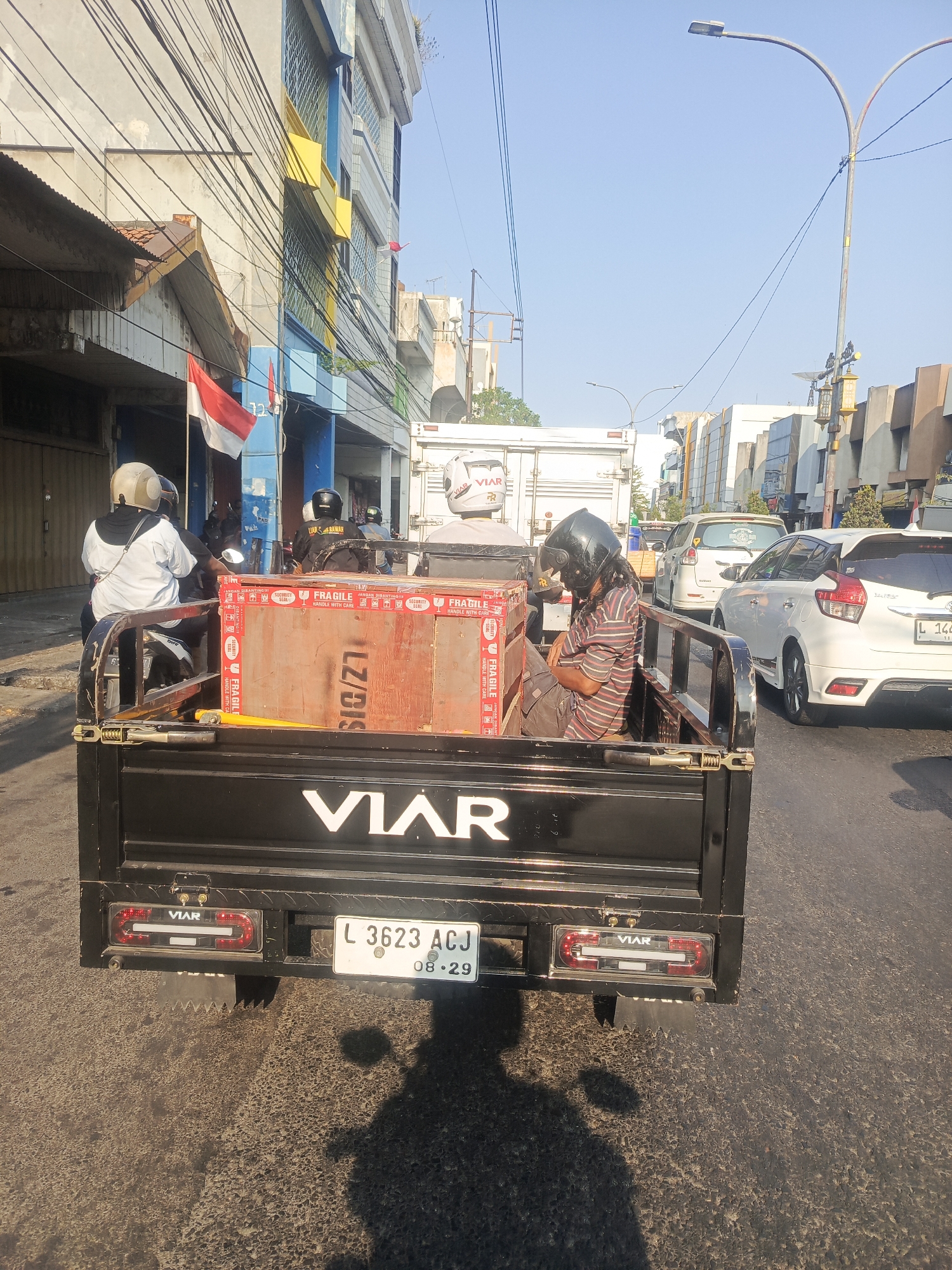 The image captures a street scene in Surabaya, Indonesia, likely on a sunny day. In the foreground, a black pickup truck with the "VIAR" logo is prominent. Inside the truck bed, a person wearing a striped shirt and dark pants is sleeping, with a dark helmet on their head. A large wooden crate with "FRAGILE" and "HANDLE WITH CARE" markings is also present in the truck bed. The truck has a license plate that reads "L 3623 ACJ 08.29". Behind the pickup truck, other vehicles are visible, including a white car and a white delivery truck. The buildings lining the street are a mix of modern and older structures, with visible electrical wires overhead. Indonesian flags are displayed on one of the buildings. The overall impression is a typical urban traffic scene in Indonesia.