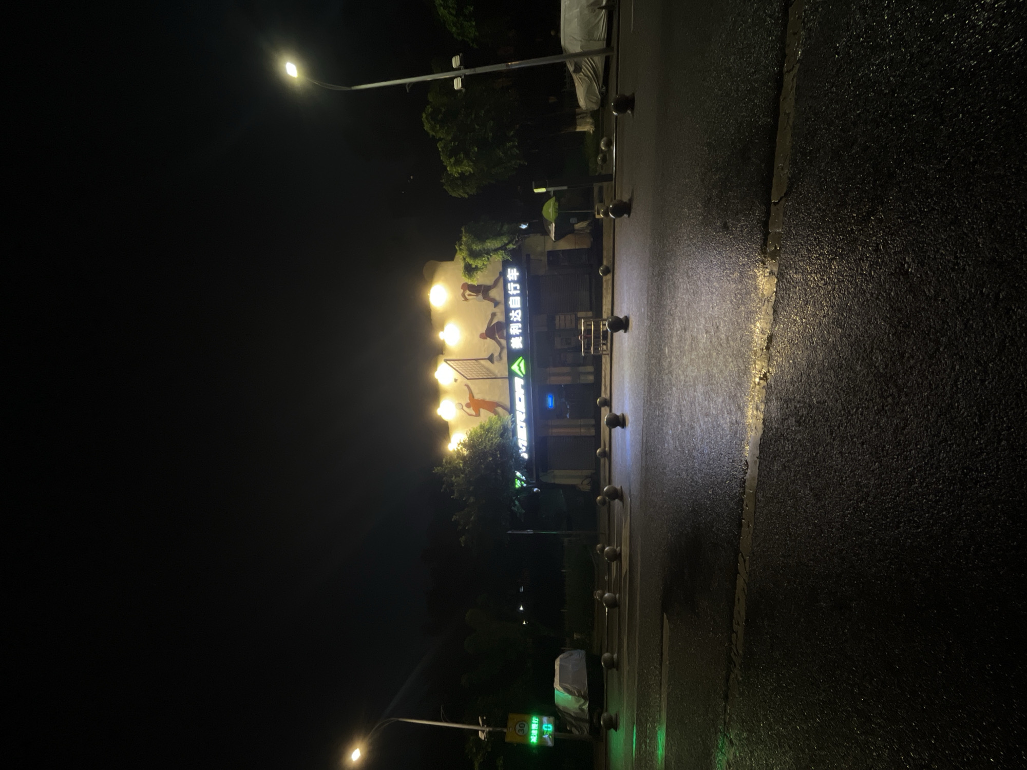 The image displays a nighttime urban street scene in Jiulongpo District, China, following a recent rainfall, indicated by the wet and reflective asphalt road that dominates the foreground. In the midground, a light-colored building features a prominent illuminated sign displaying the English word "MERIDA" alongside the Chinese characters "美利达自行车" (Merida Bicycles), identifying it as a bicycle shop. Above this sign, the building's facade is adorned with murals depicting stylized human figures engaged in sports, illuminated by several upward-facing warm-toned lights. The ground floor of the building shows closed storefronts, with visible roller shutters.
The road is dark asphalt with faint yellow lane markings and is punctuated by a series of dark, spherical bollards arranged horizontally across its width. On the left side of the frame, a streetlight pole emits a bright, warm light, casting illumination onto a traffic signal. This signal displays a green light with a numerical countdown of "0," accompanied by the Chinese characters "减速慢行" (reduce speed) and the number "30," likely indicating a speed limit. Adjacent to the left curb, a vehicle is parked, obscured by a white protective tarp. On the far right, another streetlight pole with a similar warm light is visible. Dark trees and foliage are present on both sides of the street and behind the main building. The sky above is dark and cloudless. No individuals are present or engaged in active interaction within the visible scene.
