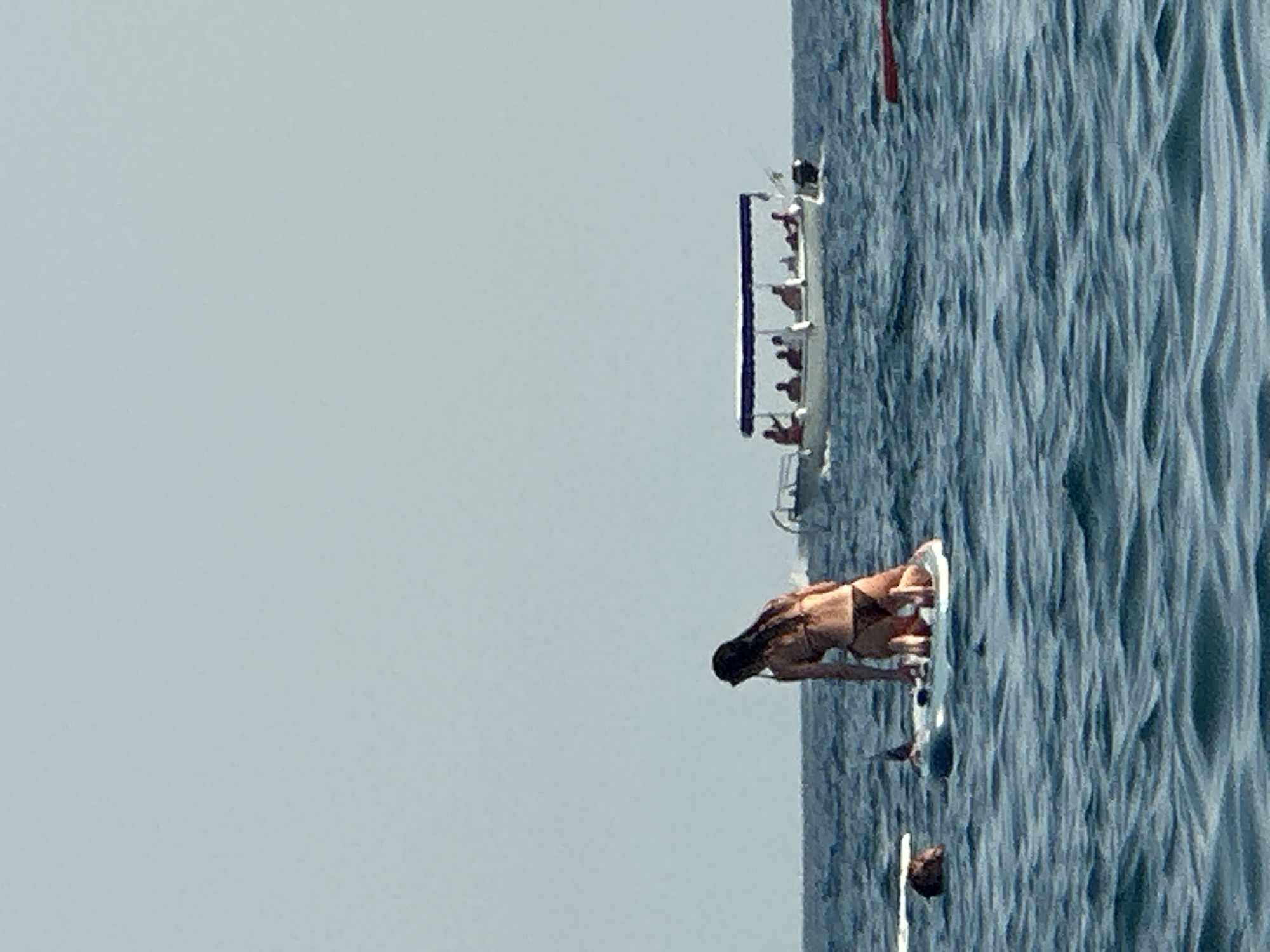 In the clear blue waters off the coast of Boreti, Montenegro, a woman is seen kneeling on a paddleboard. She is wearing a dark bikini and has her back to the camera, with her wet hair falling down her back. Her stance suggests she may be preparing to paddle or has just finished doing so. In the background, a white tour boat with a blue canopy is navigating the sea, carrying several passengers. To the left of the paddleboarder, another person is partially visible in the water, appearing to be swimming or floating. The sea is moderately choppy with visible waves. The overall scene depicts a recreational activity on the water, possibly a tour or a day out at the beach.