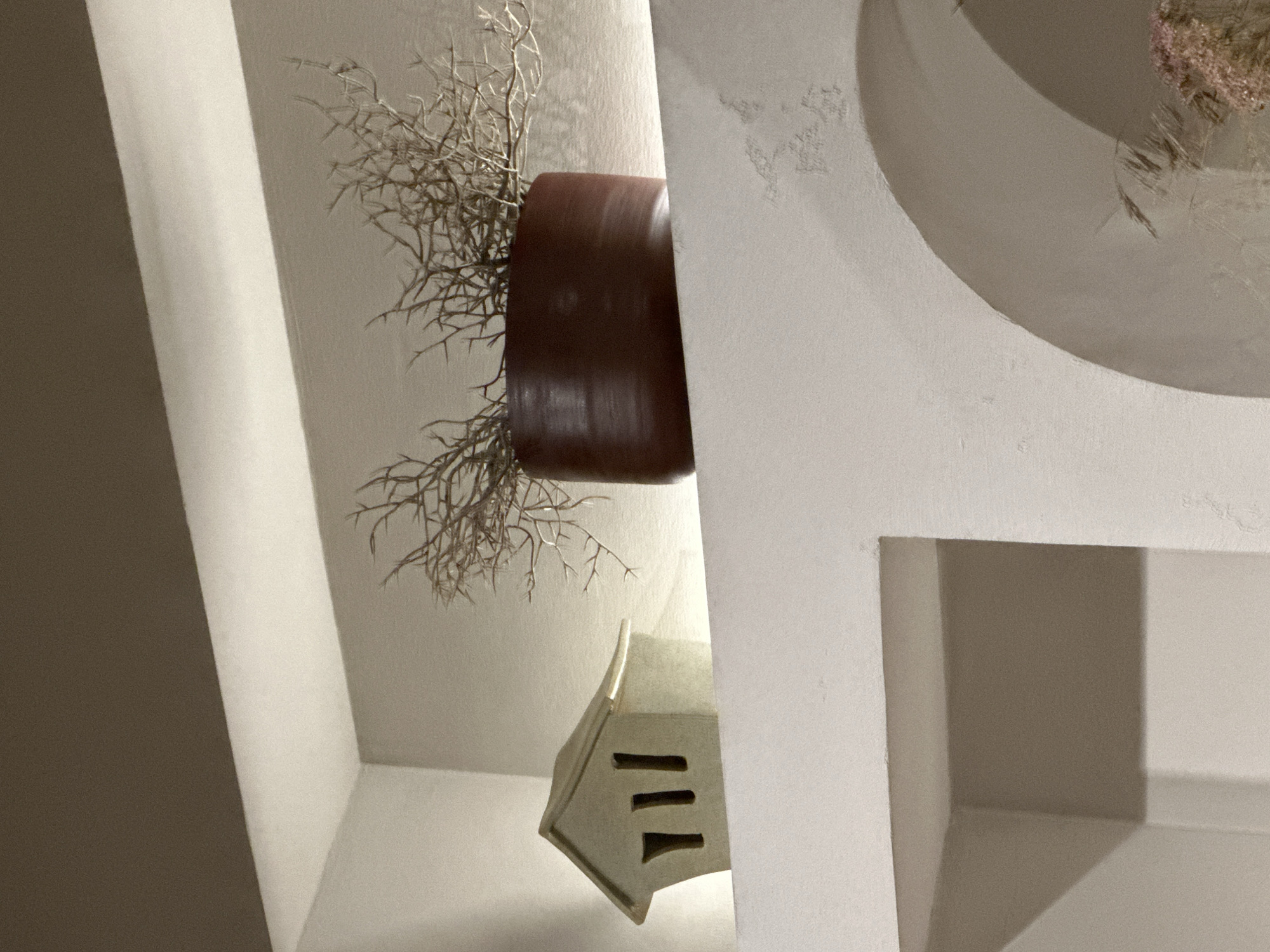 This image captures a close-up, eye-level view of a decorative shelf. On the left, a small, light-colored ceramic house sits, featuring three narrow, vertical openings on its front. To the right of the house, a dark reddish-brown ceramic pot contains dried, twig-like branches, which spread out widely. The shelf is illuminated by a soft, diffused light, creating subtle shadows and highlighting the textures of the objects and the wall. The background consists of a neutral, muted color palette of whites and grays, with a darker horizontal band across the top. The scene is static and appears to be indoors, likely in a home or commercial space. There is no indication of a specific location within Ufa, Russia; the image provides no geographical cues.