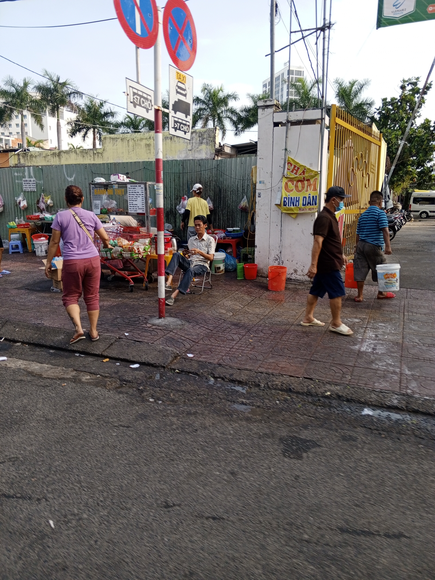 The image depicts a street scene in Nha Trang Ward, Vietnam. A woman in a purple shirt and red pants walks away from the camera towards a food stall. She is carrying a bag over her shoulder. Behind her, a food cart with a canopy is laden with various items. To the right, a man sits on a chair, looking at his phone. Further to the right, two men walk along the sidewalk. One man wears a brown shirt and blue shorts, the other wears a striped blue and white shirt and grey shorts. Both are carrying buckets. In the background, a yellow gate and a building are visible. Traffic signs indicating "TAXI" and bus routes are posted on a pole in the foreground. The ground is paved with tiles, and there is a section of asphalt road. Several orange buckets are placed along the sidewalk. The scene appears to be an outdoor market or street food area.