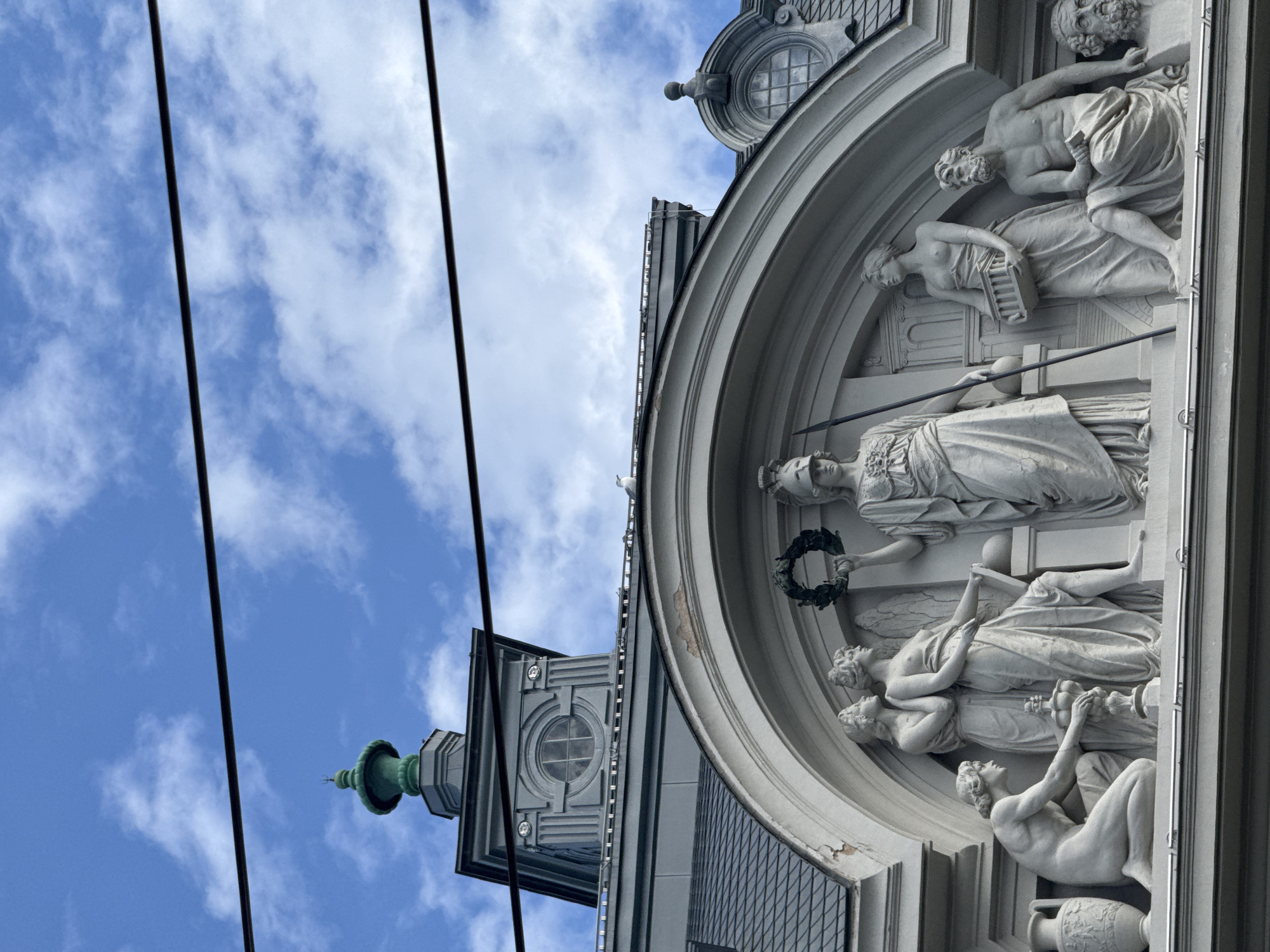 The image depicts the facade of a historical building in Riga, Latvia, featuring a semi-circular sculptural relief adorned with classical figures. The relief, likely made of white stone or plaster, showcases several human figures in draped garments, possibly representing allegorical or mythological scenes. Above the relief, there is architectural detailing, including a smaller window and cornice. To the left of the facade, a tower-like structure with a green, onion-shaped dome and decorative windows is visible. In the background, the sky is blue with scattered white clouds. Two black power lines are running across the scene. A small bird, likely a pigeon, sits atop of the arch.