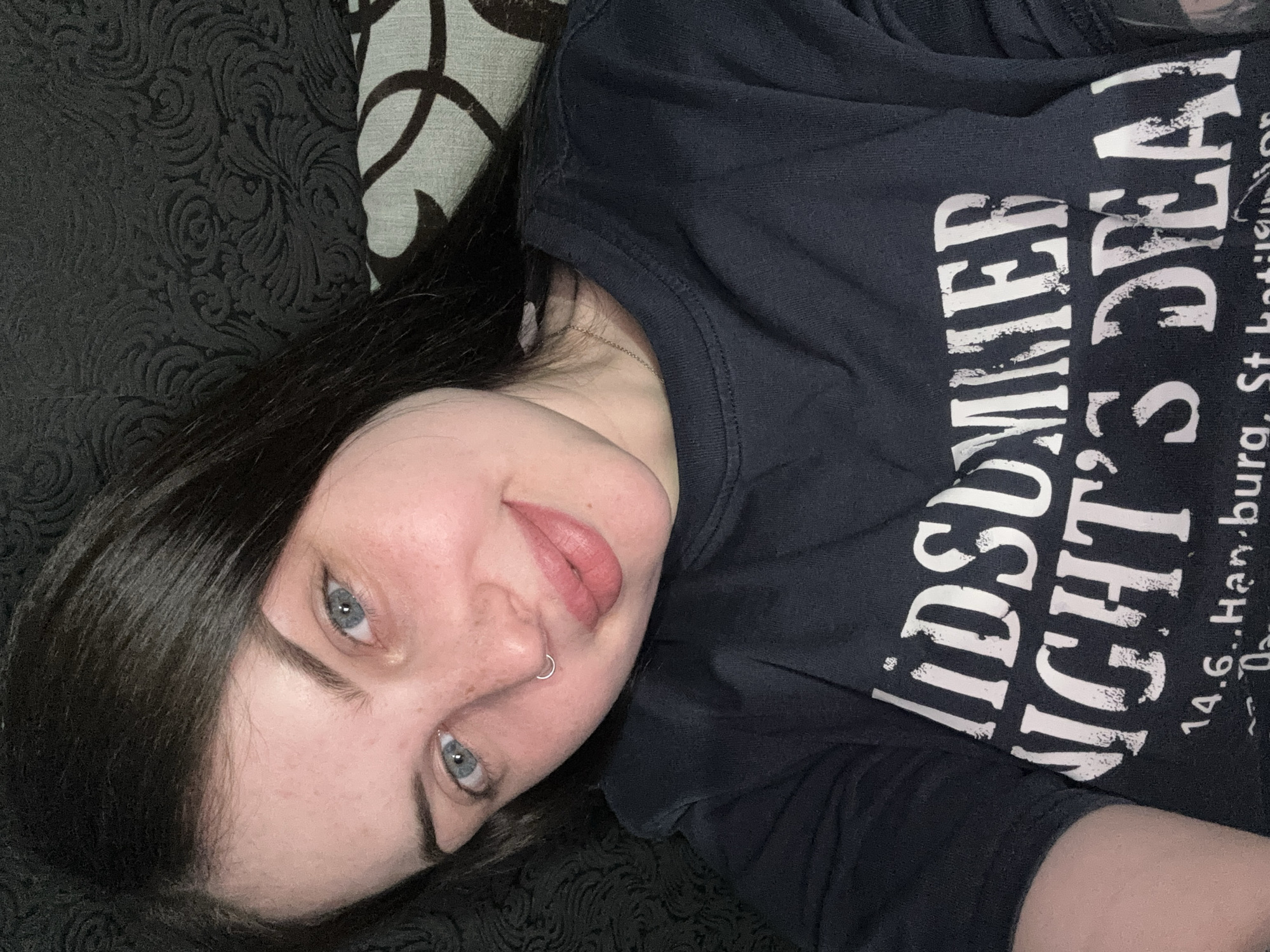 A young woman with pale skin, dark hair, and blue eyes is lying on a dark, patterned couch. She has a nose ring. She is wearing a dark-colored t-shirt with white lettering that reads "MIDSUMMER NIGHT'S DEAD". The text below the main phrase appears to include the location "14.6...Hamburg, St. Hetlinger". The background includes the dark, textured couch and a patterned pillow. The location is not specified as Chornomorskoe, Ukraine.