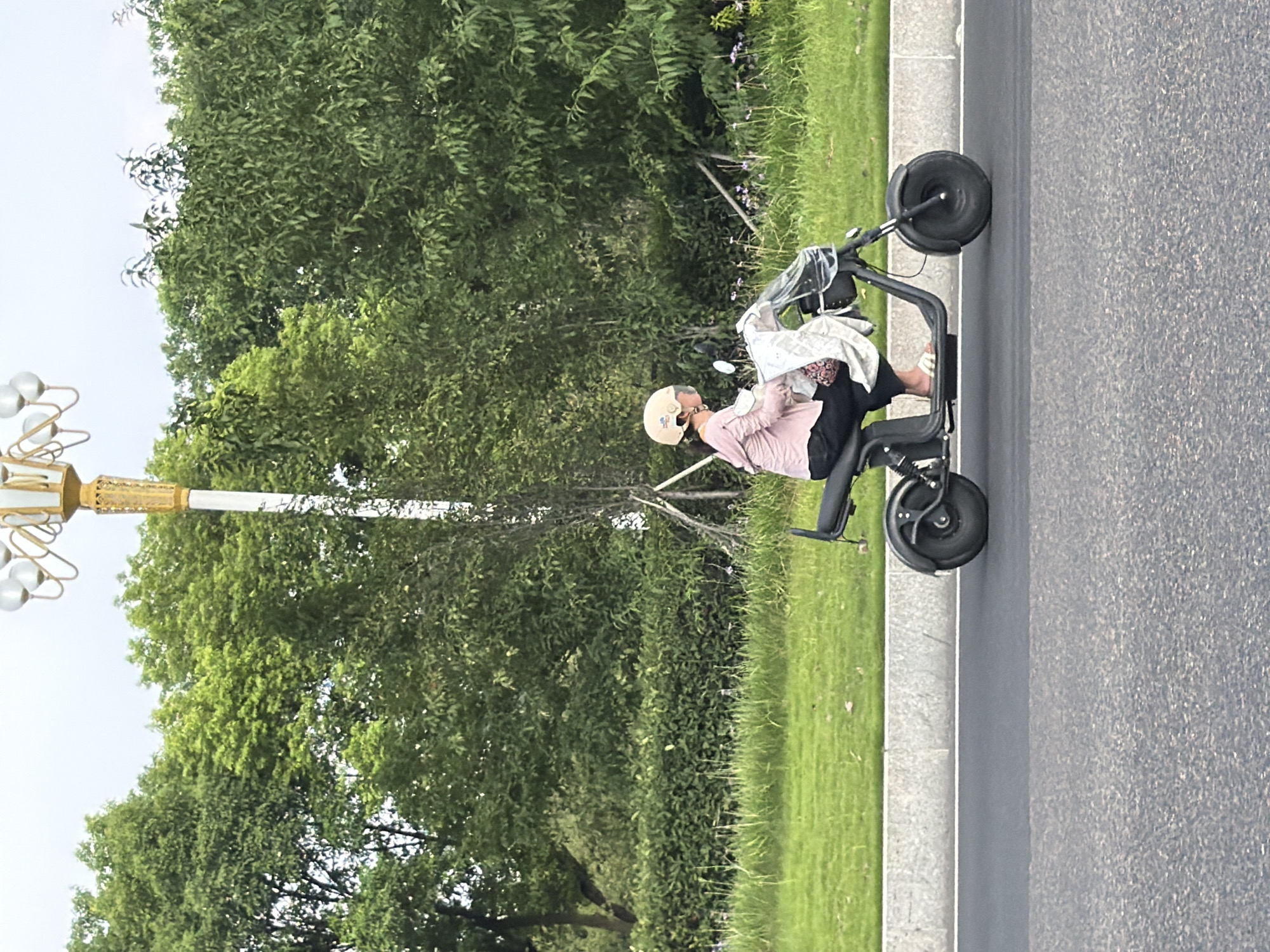 The image depicts a person riding a black electric scooter on a paved road, with lush green trees and grass lining the roadside. The rider is wearing a pink long-sleeved shirt, black pants, a light-colored helmet, and white sandals. A white cloth or bag is draped over the front of the scooter. In the background, a tall lamppost with multiple globes stands prominently against the sky. The scene appears to be in an urban or suburban area with well-maintained landscaping.
