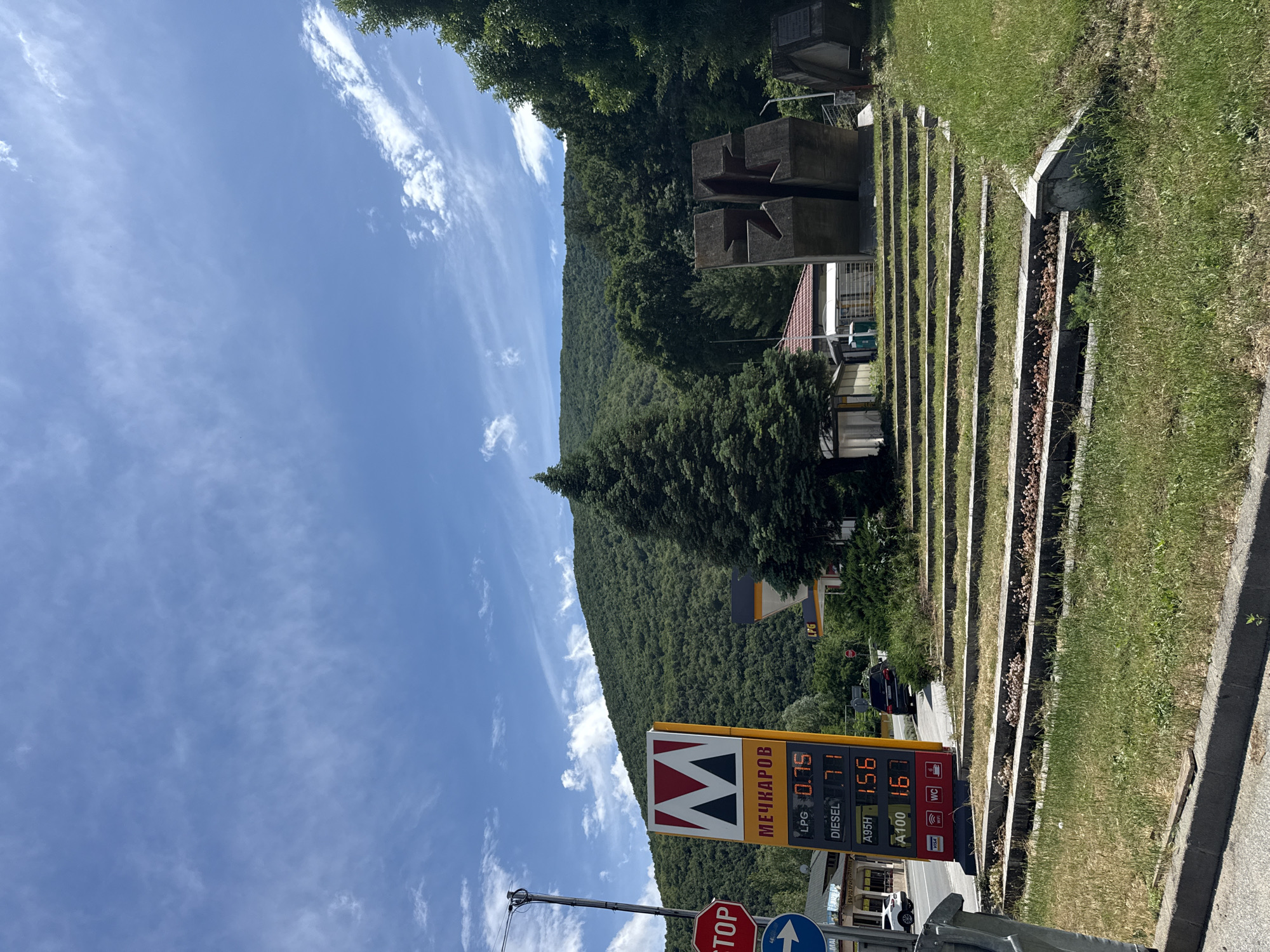 This image depicts an outdoor scene in Vetren Dol, Bulgaria, captured on a bright, sunny day with a clear blue sky dotted with wispy white clouds. The central focus is a tall, vertical gas station sign for "МЕЧКАРОВ" (Mechkarev). The sign displays current fuel prices in Cyrillic, likely in Bulgarian levs per liter: LPG at 0.75, Diesel at 1.71, A95H at 1.56, and A100 at 1.67. It also indicates availability of WC, WiFi, and acceptance of VISA payments.
In the foreground, partially obscured, are a red octagonal STOP sign and a blue circular directional sign with a white arrow. To the right, the ground slopes upwards with neatly arranged concrete terraces, filled with grass and some sparse vegetation. In the middle ground, substantial concrete structures, possibly part of a monument or architectural installation, are visible among dense trees. A building with a reddish-brown tiled roof can be seen behind these structures.
The background is dominated by a vast, rolling mountain range covered entirely in lush, dense green forest, extending to the horizon under the clear sky. The overall scene suggests a blend of natural landscape and roadside amenities in a mountainous region, under pleasant weather conditions.