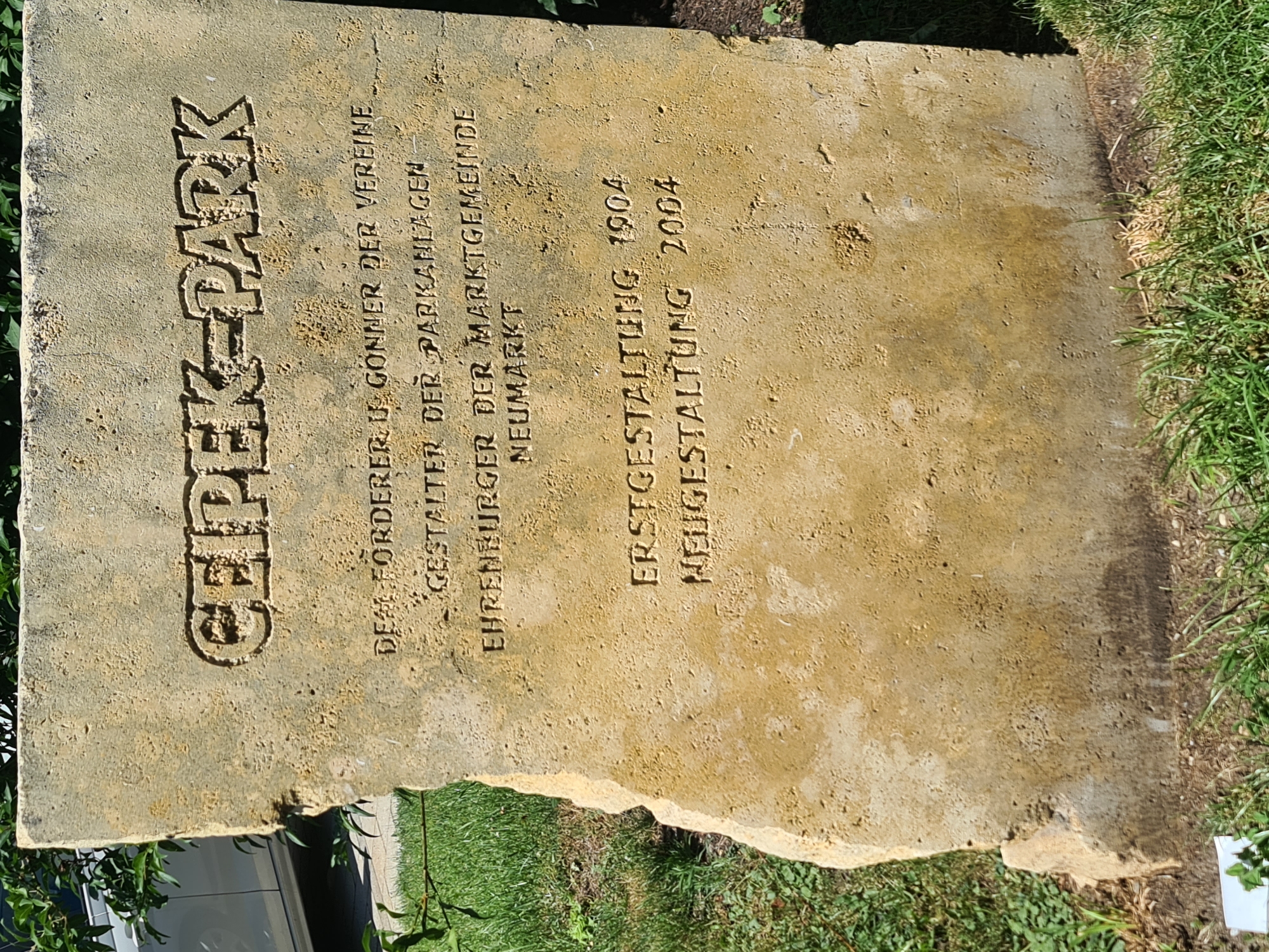 A weathered, light-colored stone monument stands upright in a grassy area with some green foliage and a portion of a white car visible in the background. The monument has an irregular shape with a chipped left edge. Engraved text appears on the front of the stone. The top line is in large, bold, sans-serif capital letters reading "GEIPEK-PARK". Below this, smaller engraved text lists acknowledgements and dates. The text reads: "DEM FÖRDERER U. GÖNNER DER VEREINE GESTALTER DER PARKANLAGEN EHRENBURGER DER MARKTGEMEINDE NEUMARKT ERSTGESTALTUNG 1904 NEUGESTALTUNG 2004". The monument is situated outdoors, likely in a park or public space. The lighting suggests it is daytime, with the sun casting shadows and highlighting the textures of the stone.