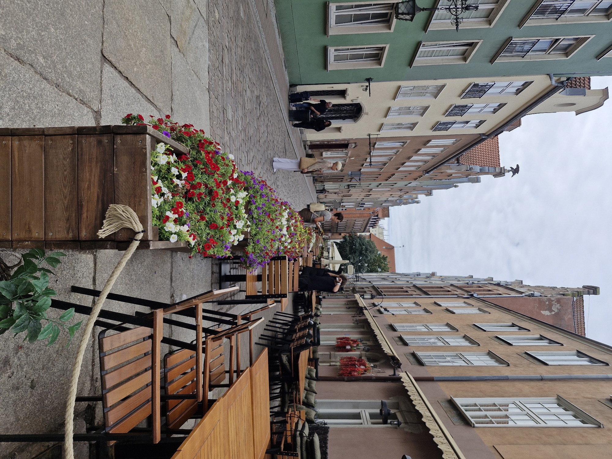 This image depicts a street scene in the city of Gdańsk, Poland, likely within the historic Old Town, given the architecture and cobblestone street. The perspective is looking down a narrow street lined with colorful, multi-story buildings in various shades of green, cream, and brown.
In the foreground, a large wooden planter box filled with vibrant red, white, and purple flowers is prominently displayed. A thick rope barrier is strung in front of the planter, with a small green plant visible in the lower right corner. The street itself is paved with large, irregular cobblestones.
Several tables and chairs from outdoor cafes are arranged along the right side of the street, with wooden tabletops and chairs with black metal frames. Some tables are set with place settings, and awnings extend from the buildings overhead.
People are present in the scene. On the left side of the street, two individuals, one in black and another in a dark hooded sweatshirt, are near an entryway, with a black suitcase beside them. Further down the street, a woman in a light-colored dress and a straw hat stands near the flower planter, holding a woven bag. On the right side, a person in black is taking a photo with a phone, standing behind some of the cafe tables. Another woman in a striped shirt is visible further down the street.
The architecture of the buildings is characteristic of historic European cities, with tall, narrow facades, numerous windows, and ornate detailing on some of the rooftops. The sky is overcast with a pale, cloudy appearance. The overall impression is a lively street scene with a blend of urban architecture, natural elements, and human activity.
