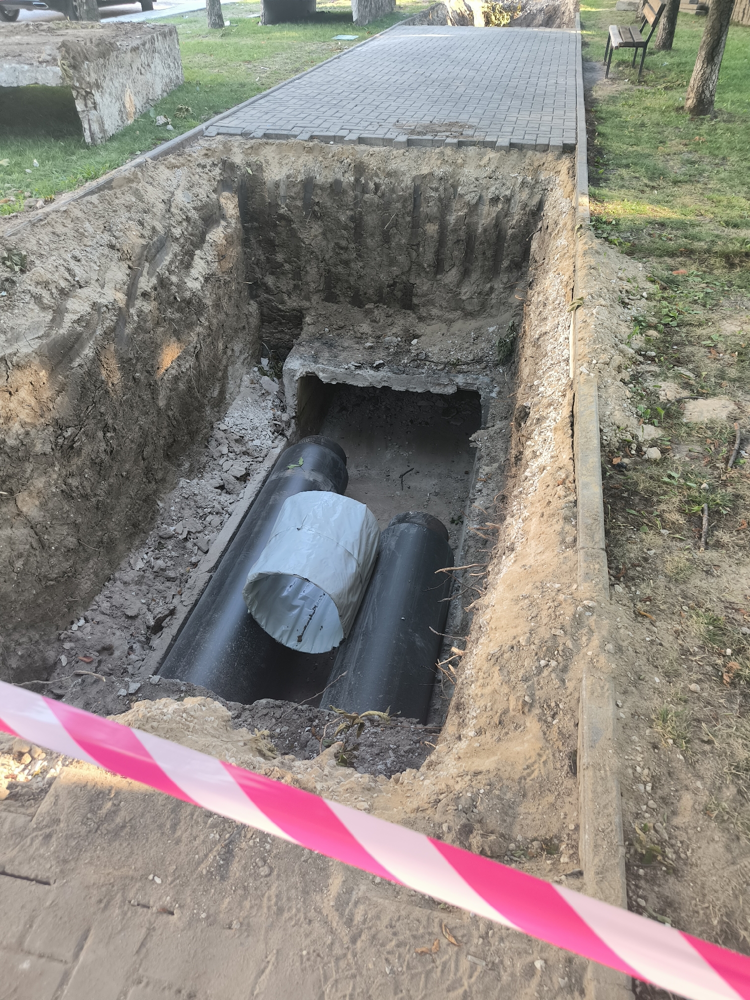 An aerial view captures a trench dug into the ground, revealing large black pipes. The trench is situated next to a paved walkway and a grassy area. A pink and white striped caution tape stretches across the foreground, indicating a restricted zone. The scene appears to be an urban setting, possibly a park or sidewalk in Chișinău, Moldova, where infrastructure work is underway. The excavation is deep, exposing layers of soil and some debris. There are no people visible in the image.