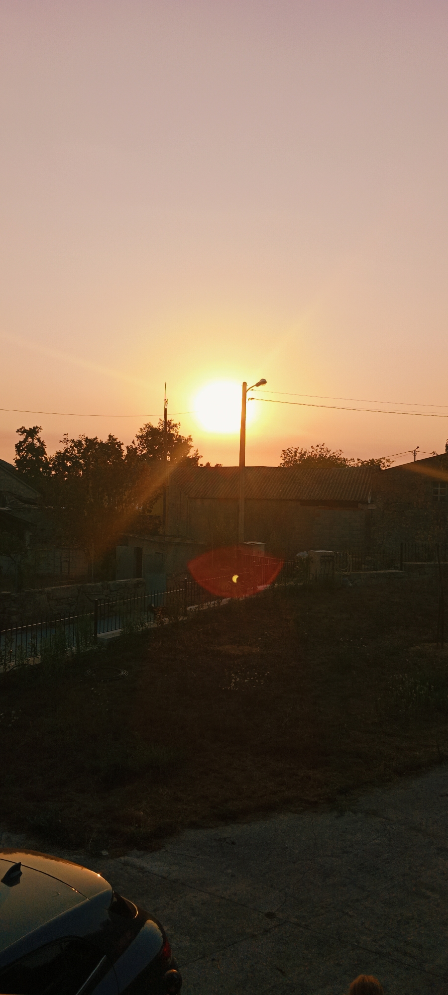 The image captures a sunset over a residential area. The sky is a gradient of pale pink and orange, with the sun a bright, intense orb radiating light. A wooden utility pole with a streetlamp stands prominently, silhouetted against the sky. Trees, also dark silhouettes, are scattered across the middle ground, with rooftops of houses visible behind them. A metal fence borders a raised grassy area in the foreground, which slopes down to a paved surface where the back of a dark-colored car is visible. The overall scene suggests an evening in a quiet, possibly rural or suburban, part of Adai, Spain.