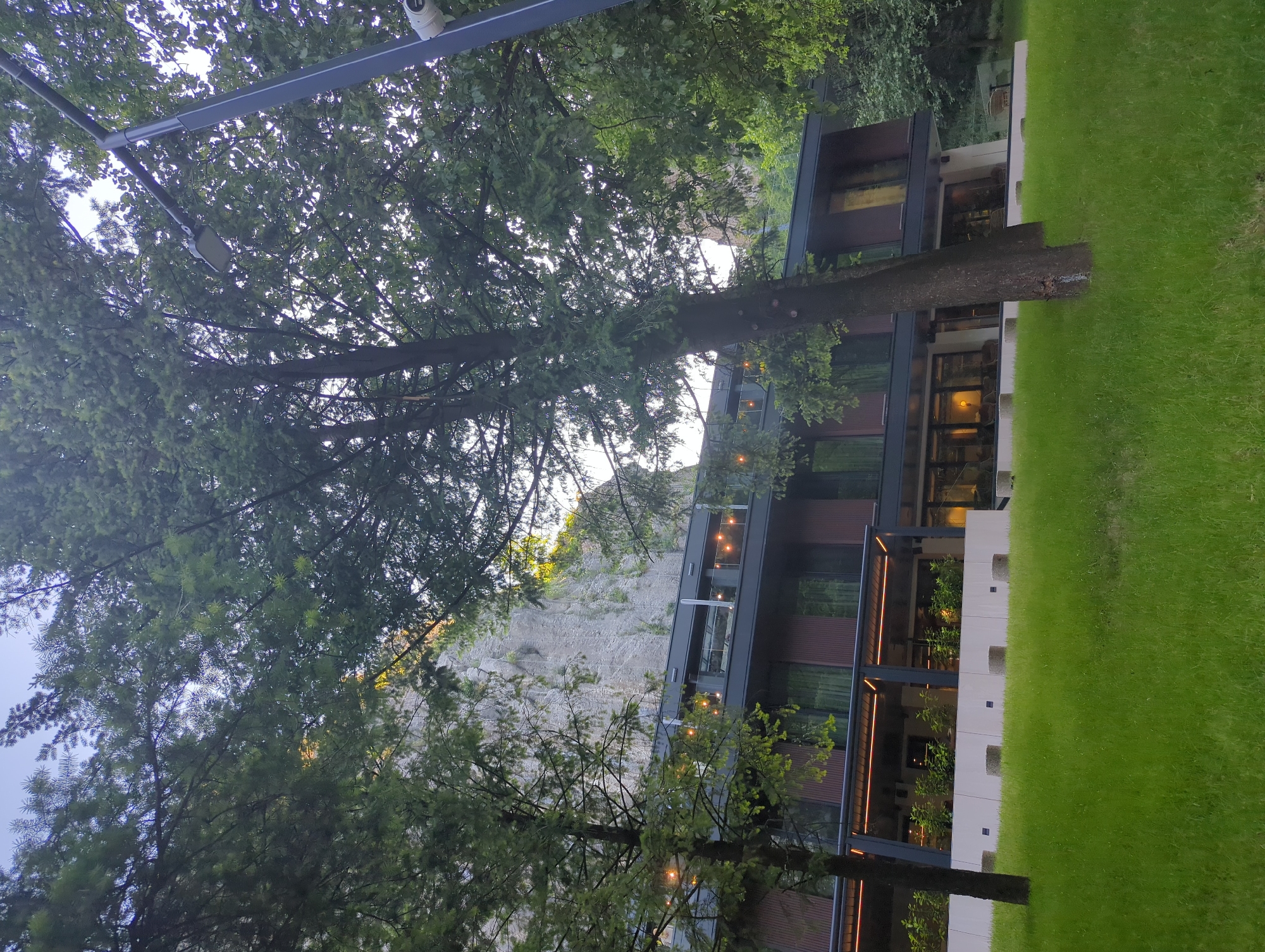 The image captures a modern building nestled amidst lush greenery, likely a hotel or a resort. The architecture features clean lines, large glass windows, and dark-colored siding, suggesting a contemporary design. The building is surrounded by well-maintained green grass on one side and dense trees on the other. A large tree trunk is prominently visible in the foreground, partially obscuring the building.
The scene appears to be set during dusk or early evening, as evidenced by the warm glow of lights emanating from within the building, visible through the windows and along the balconies. The lighting creates an inviting and serene atmosphere. The sky is overcast or the light is muted, contributing to the twilight ambiance.
In the background, a steep, rocky incline or cliff face is visible, partially covered by vegetation. The presence of trees, the cliff, and the manicured lawn suggest a natural or semi-natural setting, possibly in a mountainous or hilly region. There are no visible people in the image, and the focus is on the building and its surrounding environment. A security camera is mounted on a pole above the trees on the left side of the image. The overall mood is one of tranquility and a harmonious blend of modern architecture with nature.