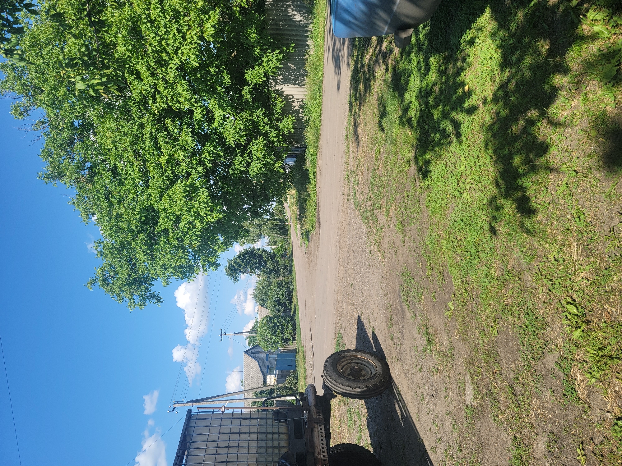A sunny day in Vovkivka, Ukraine, showcases a rural scene with a tractor in the foreground. The tractor, with its large tire clearly visible, is parked on a dirt and grass area beside a gravel road. In the background, a line of houses with grey roofs and blue fences line the road, interspersed with trees and foliage. A large, leafy green tree dominates the right side of the frame, casting dappled shadows on the grass. The sky is a bright blue with scattered white clouds. The overall impression is one of a quiet, agricultural community.