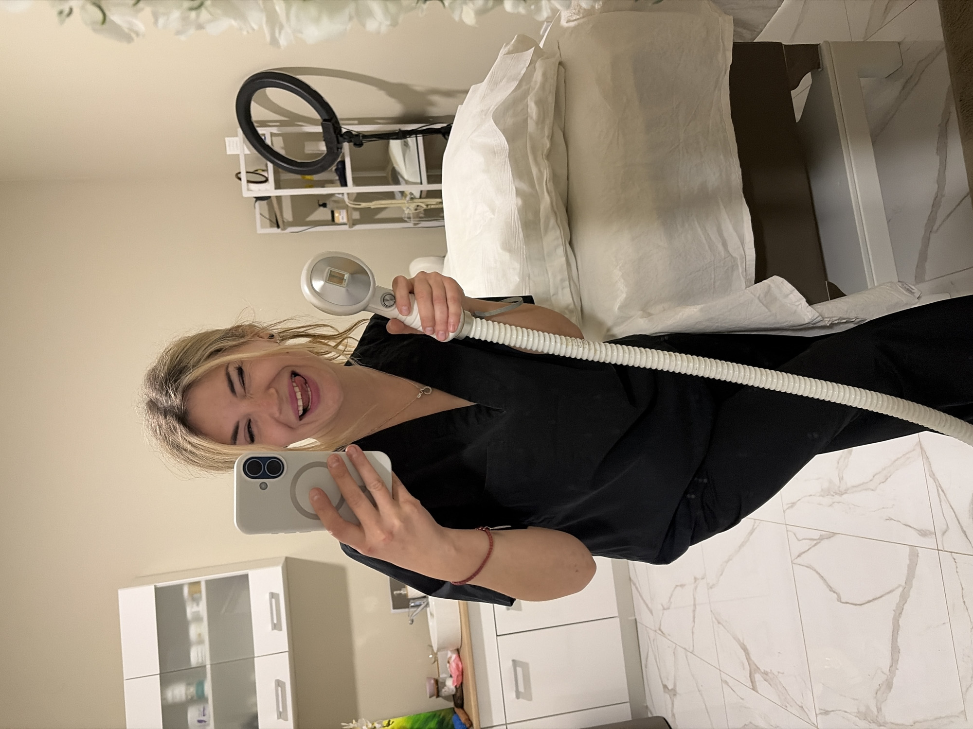 A woman with blonde hair, wearing a black scrub top, is captured in a selfie taken in a professional setting, likely a beauty salon or clinic in Budva, Montenegro. She holds a white smartphone with a visible MagSafe ring in her left hand, using it to take the photo. In her right hand, she grasps a white, futuristic-looking device connected to a long, corrugated white hose, possibly an apparatus for a beauty or medical treatment.
The woman is smiling broadly, revealing her teeth and tongue, conveying a joyful and friendly demeanor. The background features a clean, modern interior with light-colored walls and white, marble-patterned floor tiles. To the right, a white treatment bed or table with white bedding is visible. Above it, a black ring light on a stand suggests professional lighting equipment. Further left, a white cabinet with glass doors and shelves can be seen, likely used for storage of supplies. The overall environment appears well-maintained and professionally equipped. No visible text is present in the image.