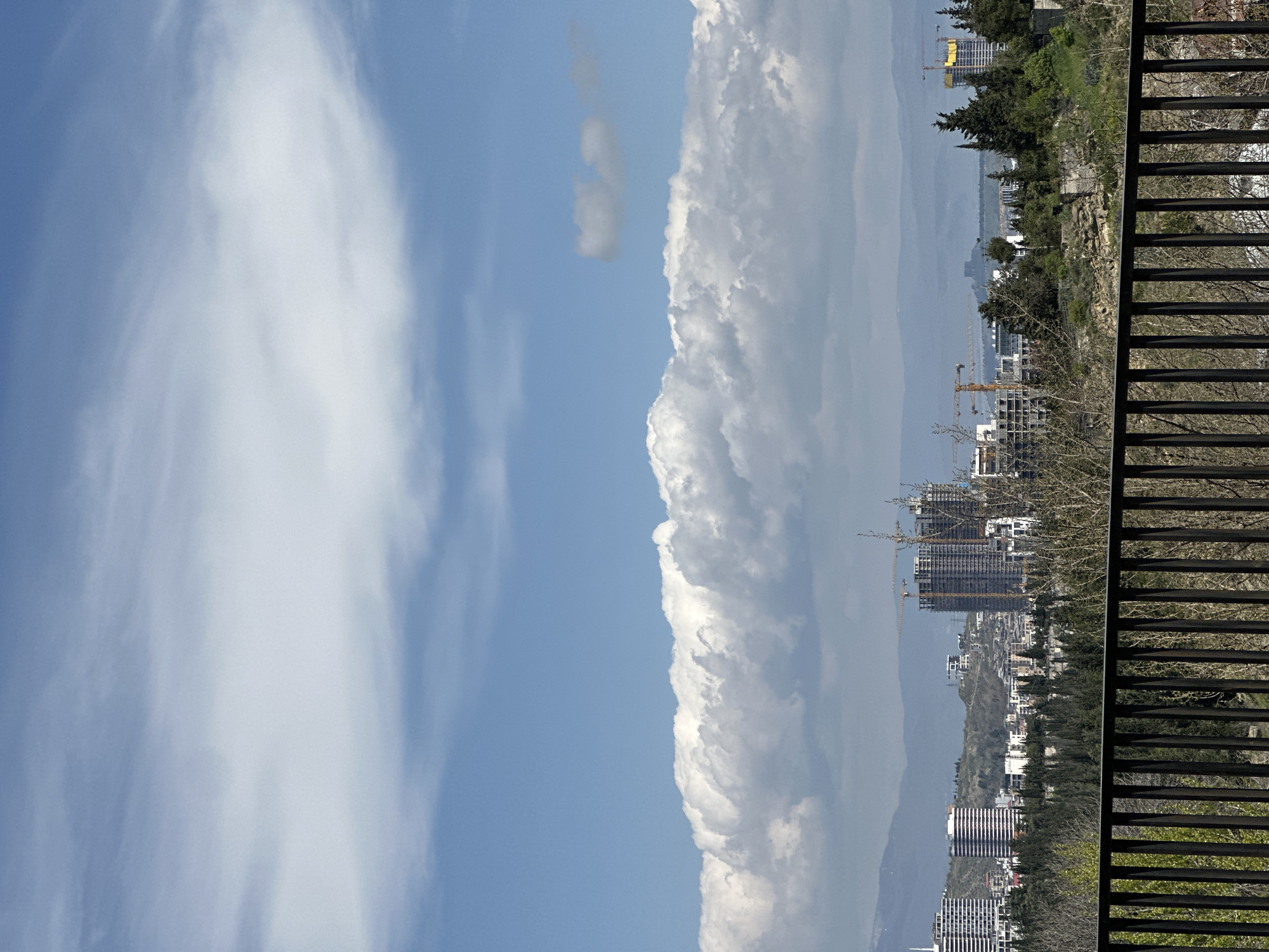 The image presents a wide, elevated view of a city under a bright sky. In the immediate foreground, a dark, slatted fence or railing runs horizontally across the bottom right, partially obscured by some bare branches and sparse green foliage.
Beyond the foreground, a sprawling urban landscape extends into the midground. Numerous multi-story buildings populate the area, with several prominent structures clearly under construction, indicated by visible scaffolding and multiple tower cranes extending above them. The city is interspersed with green trees and bushes, suggesting a mix of urban development and natural elements.
In the far distance, a chain of hazy, bluish mountains forms the horizon line, creating a soft backdrop. The sky occupies a significant portion of the upper frame, displaying a vibrant blue. A large, dense formation of white, fluffy cumulus clouds dominates the right side of the sky, appearing like a substantial cloud wall. On the left side, lighter, more wispy clouds are scattered.
The scene depicts daytime conditions, characterized by bright, even lighting and fair weather. There are no people visible, and the primary activity suggested is ongoing urban development. While no text is visible in the image, the context provided identifies the location as Tbilisi, Georgia.