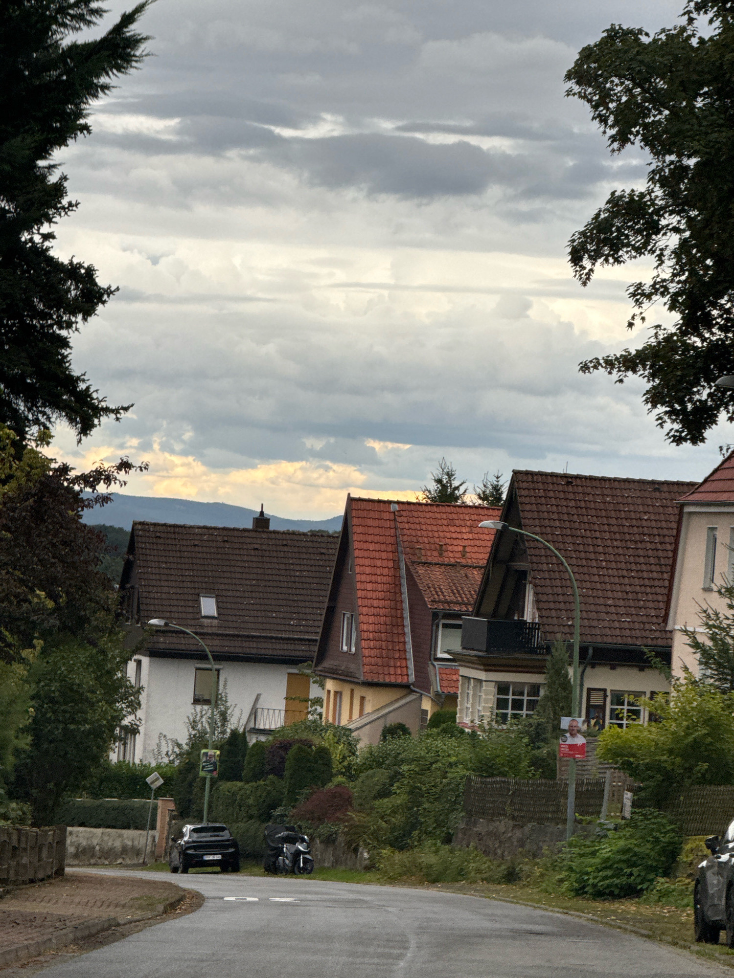 The image depicts a street in Bad Sachsa, Germany, with houses lining one side. The sky is overcast with dramatic clouds. In the foreground, a paved road curves to the left. A black SUV is parked on the right side of the road, and a motorcycle is parked behind it. Further down the street, a street lamp with a poster attached to it stands near a fence. Several houses with red-tiled roofs are visible behind the fence and surrounding greenery. Trees frame the top of the image, with one on the left and branches on the right. In the background, a distant mountain range is visible beneath the cloudy sky. The overall scene suggests a residential area under a moody sky.