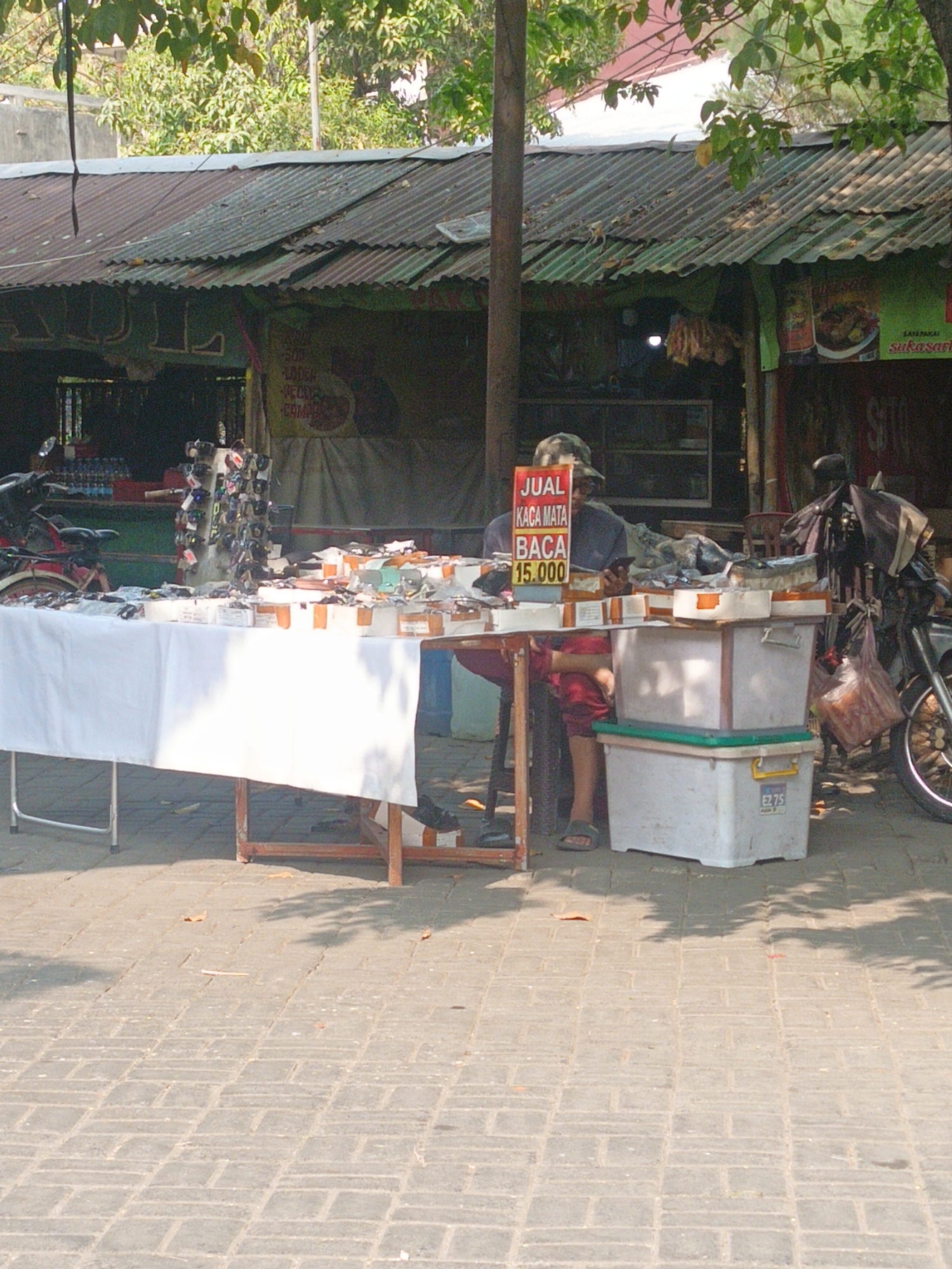 A street vendor in Wedi, Indonesia, sits behind a table covered with a white cloth, displaying various merchandise. The vendor, wearing a camouflage hat and red shorts, is holding a mobile phone. On the table are several boxes, and to the left, a display rack holds many pairs of sunglasses. Behind the table, a motorcycle is parked. The scene appears to be outdoors, with a corrugated metal roof structure providing shade and a backdrop of trees and foliage. The ground is paved with interlocking bricks. A red and white sign on the table reads "JUAL KACA MATA BACA 15.000," indicating that reading glasses are for sale for 15,000 Indonesian Rupiah.