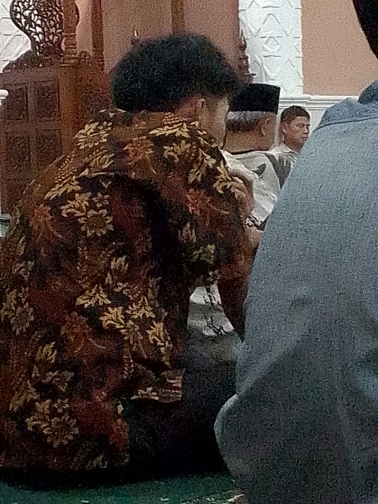 The image shows a rear view of a person wearing a dark brown, ornate patterned batik shirt. The person has dark, curly hair. To the left of this person, partially visible, is a decorative wooden structure, possibly part of an ornate pillar or screen. Behind the person in the batik shirt, a man wearing a black peci (a brimless cap) is visible, facing to the right. Further back, another man, also facing to the right, is seen with short hair. The setting appears to be indoors, with textured white wall panels and a patterned architectural element on the right. The overall scene suggests a religious or cultural gathering, likely in Kertajaya, Indonesia, given the presence of traditional attire and architectural styles. The foreground is dimly lit, with hints of a green surface, possibly a carpet or mat.