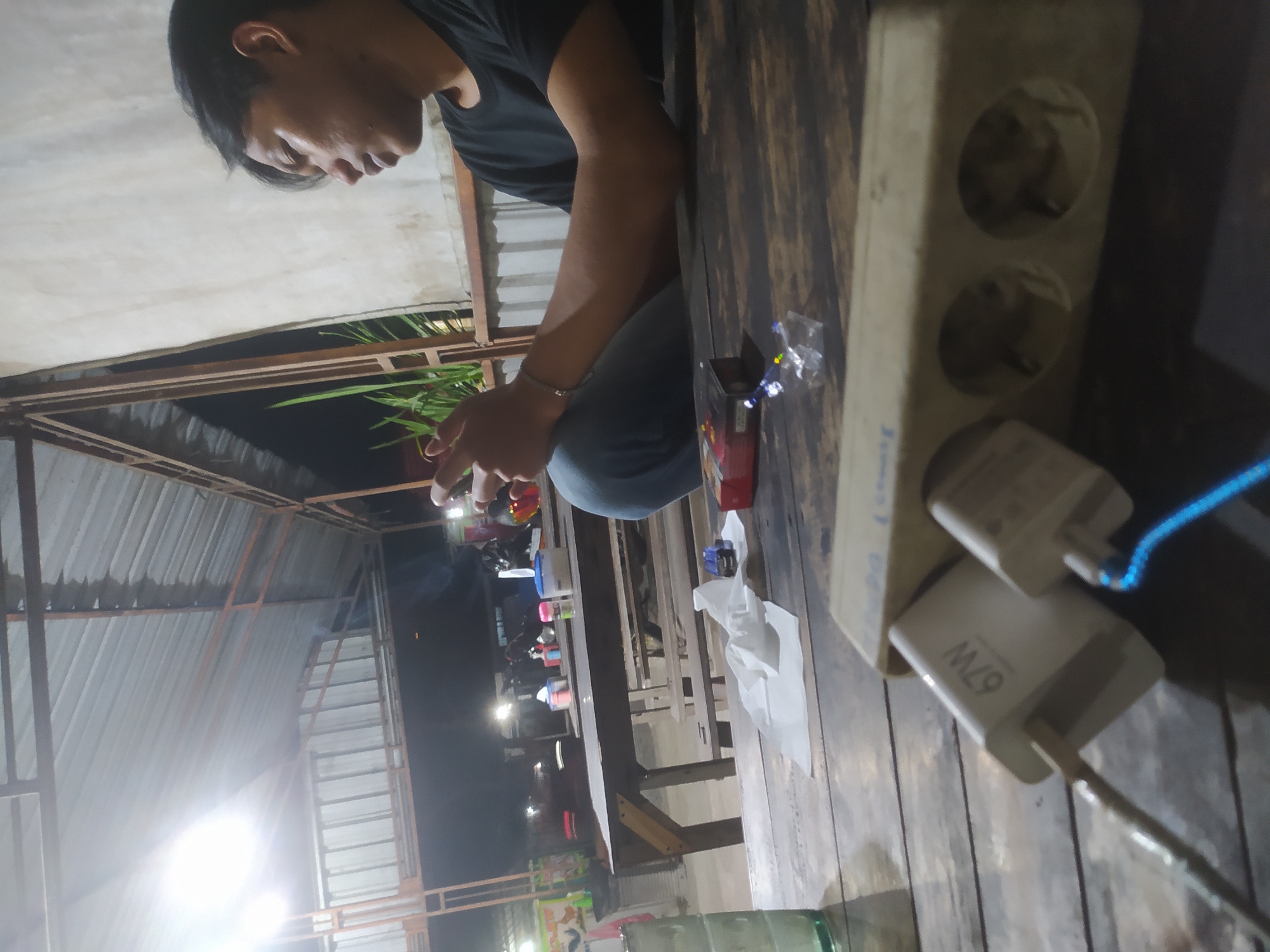 The image depicts an outdoor seating area at night, likely a street food stall or small restaurant in Tuntang, Indonesia. A young man with dark hair is seated at a wooden table, looking down at his phone. He is wearing a black t-shirt and blue jeans. The table is cluttered with various objects, including a glass, a pack of cigarettes, crumpled tissues, a small toy truck, and a power strip with two phone chargers plugged in. One charger displays "67W". In the background, there are more tables and chairs, a corrugated metal roof supported by metal beams, and some greenery. The lighting appears to be artificial, with overhead lights illuminating the scene. The overall atmosphere is casual and relaxed.