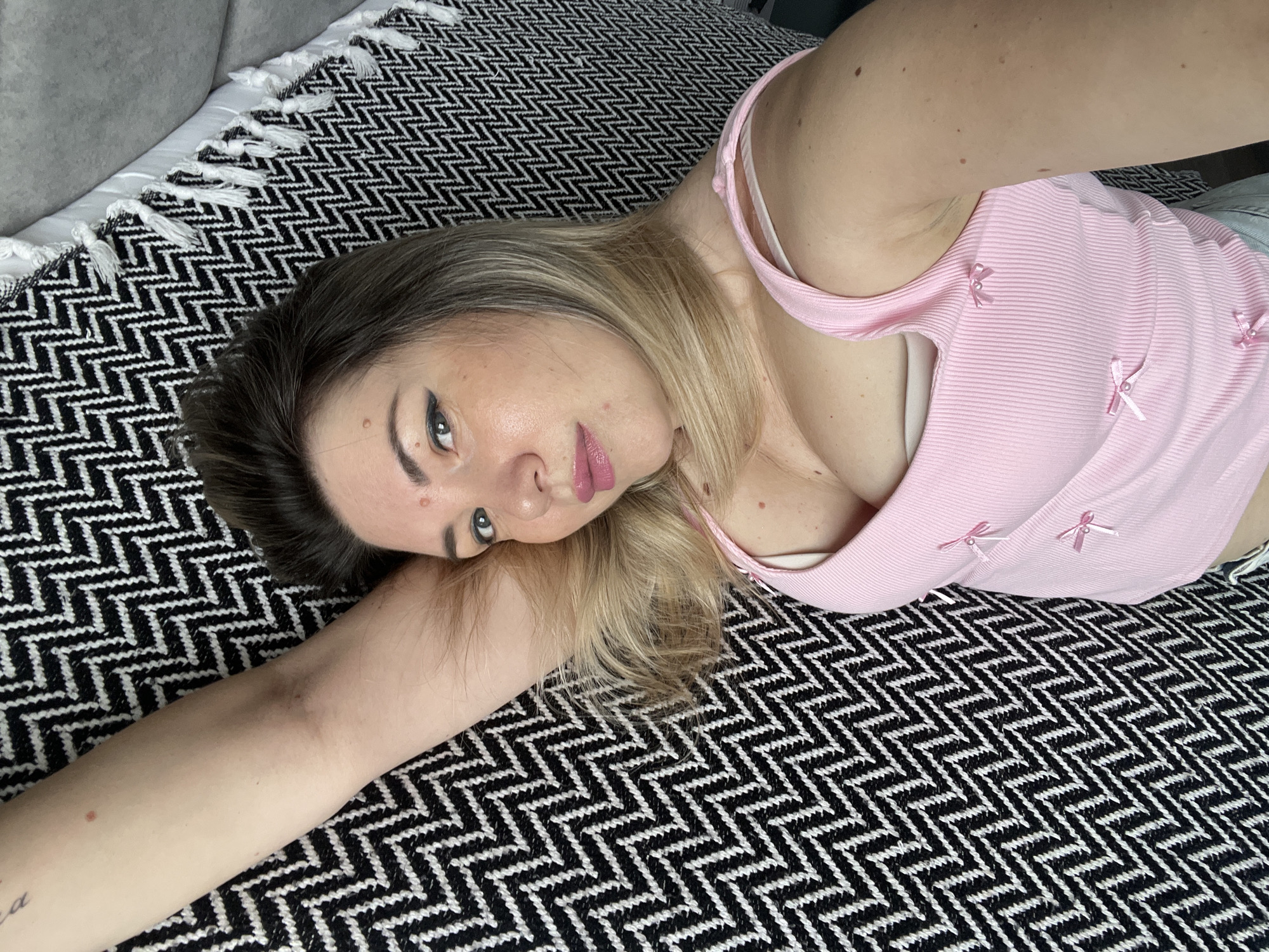 The image shows a young woman lying on a bed with a black and white patterned blanket. She is wearing a pink top. The room appears to be a bedroom. There is no discernible location in the city of Sochi, Russia.
