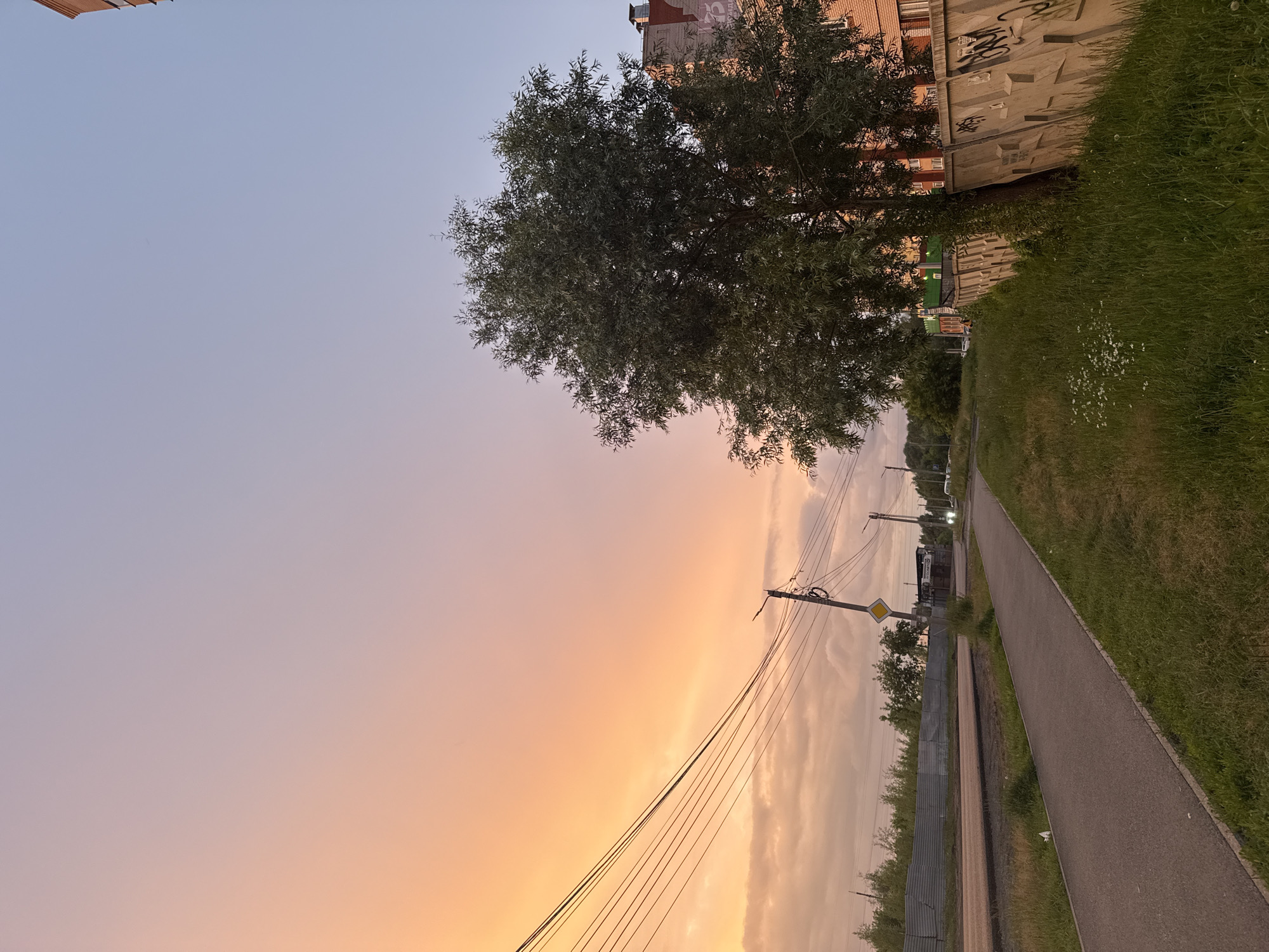 This media file depicts an outdoor scene in Izhevsk, Russia, likely during a sunset. The sky displays a gradient of soft blues and warm oranges, indicating the end of the day. A large tree with dense foliage dominates the left side of the frame, its branches reaching towards the colorful sky. Behind the tree, a multi-story building with brickwork and visible graffiti stands.
To the right of the tree, a paved pathway runs parallel to a grassy area. Along the pathway, utility poles with numerous wires stretch across the scene, suggesting an urban or suburban environment. A yellow diamond-shaped sign is visible on one of the poles. Further down the path, there are hints of buildings and vehicles, including what appears to be a small kiosk or shop. The overall mood is calm and serene, with the warm hues of the sunset creating a peaceful atmosphere. The scene is devoid of people, focusing on the landscape and the interplay of light and shadow.