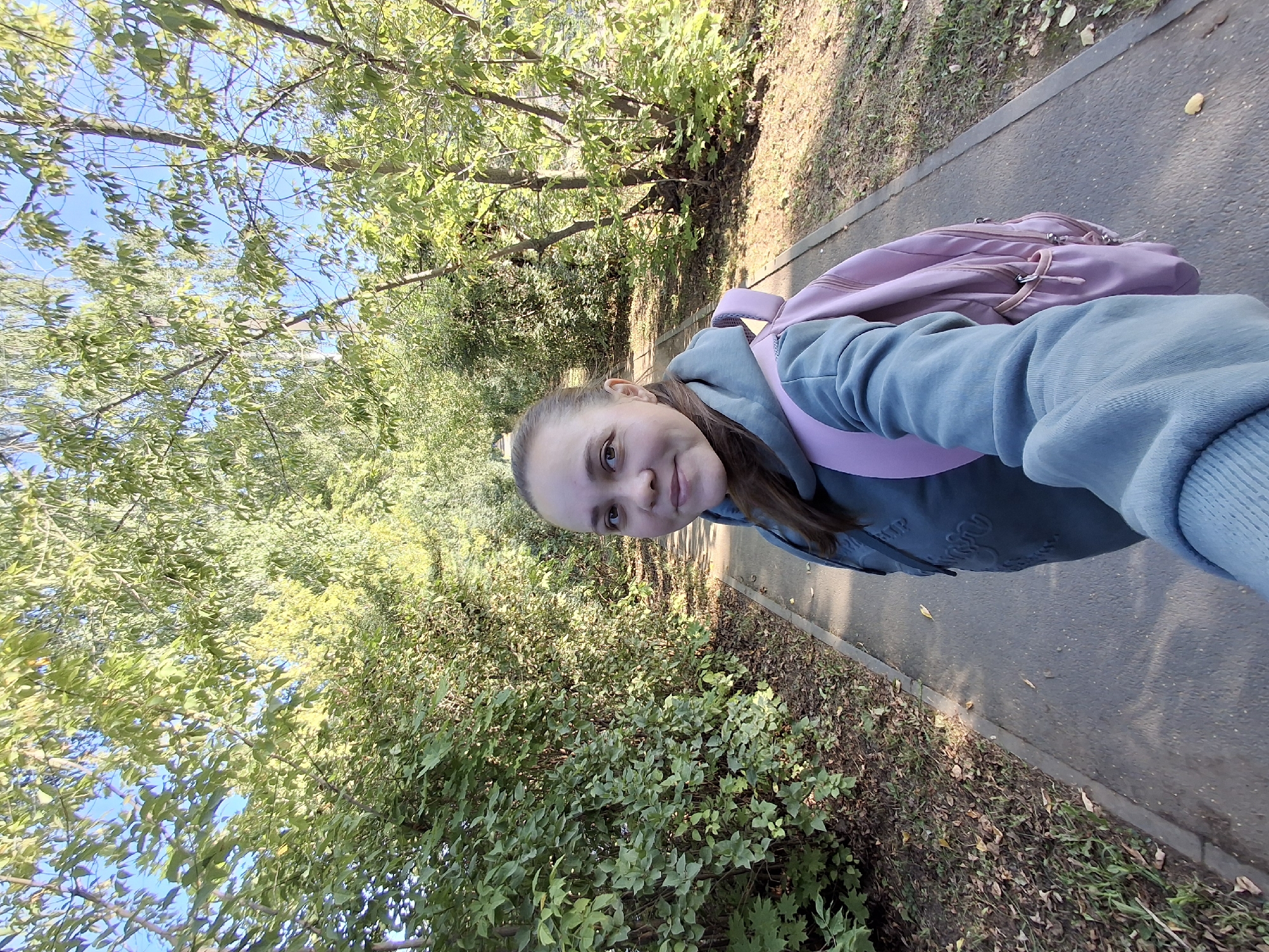 A young woman with brown hair pulled back in a ponytail is taking a selfie in a park. She is wearing a blue hooded sweatshirt and a pink backpack. The woman is standing on a paved path, with trees and foliage on either side. The sun is shining, casting shadows on the path. The location is identified as Elovo, Russia.