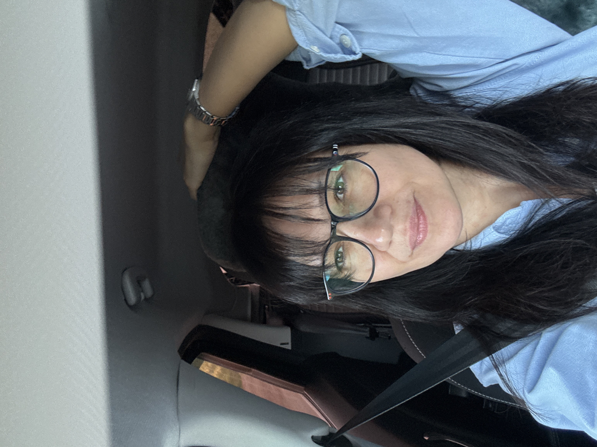 A woman with black hair and glasses is wearing a light blue collared shirt and a watch, sitting in the driver's seat of a car with the seatbelt fastened. The scene appears to be inside the vehicle in an urban environment.