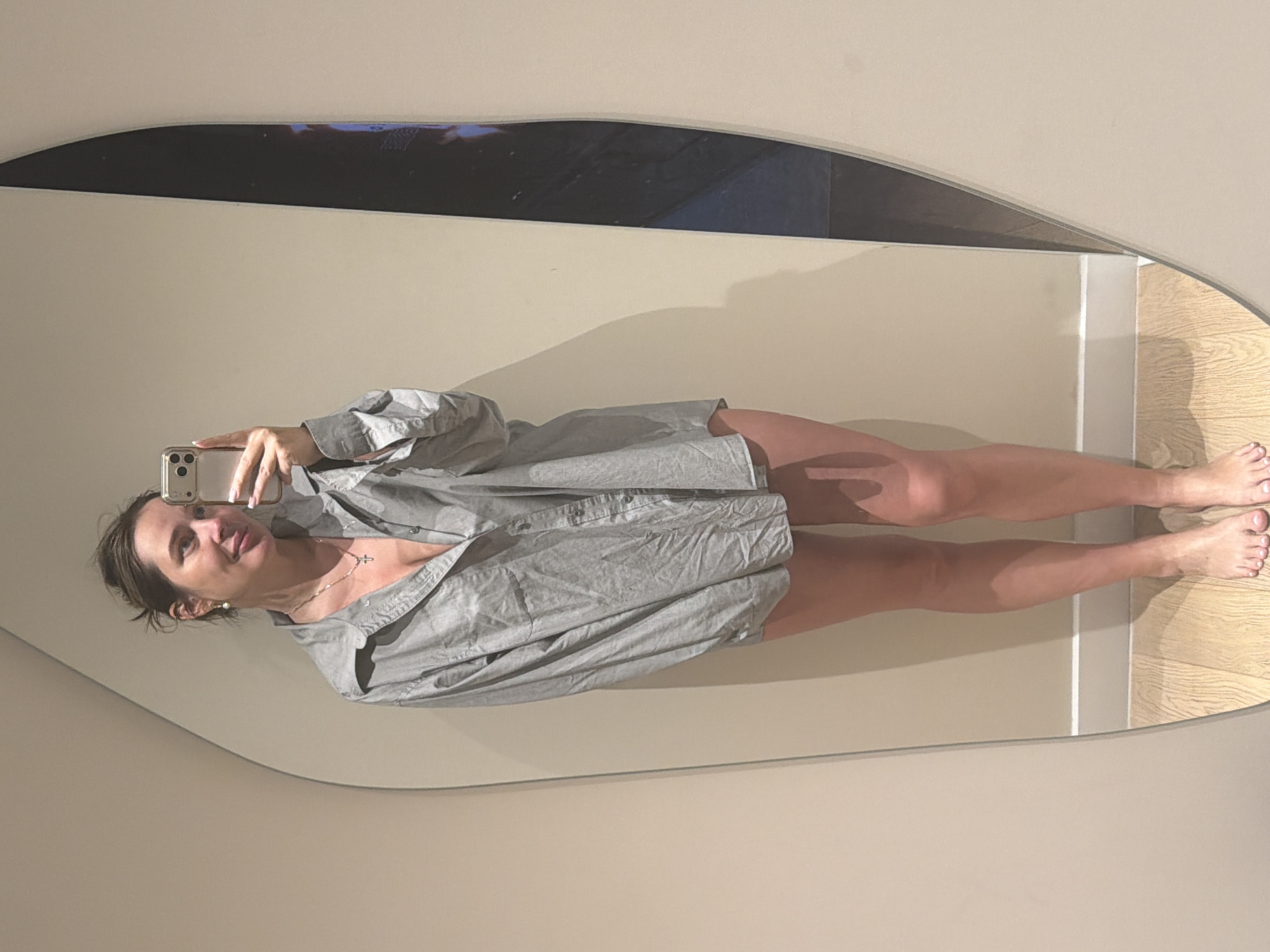 A woman is standing in front of a mirror, taking a selfie with her phone. She is wearing an oversized grey button-down shirt, which is open at the top, revealing her chest. She has on a necklace with a cross pendant and earrings. Her hair is pulled up in a messy bun. Her legs are bare, and she is standing on a wooden floor. The mirror is large and has an irregular shape. The background appears to be a wall. This image was taken indoors, in what appears to be a domestic setting. There is no indication of the location being in Moscow, Russia.