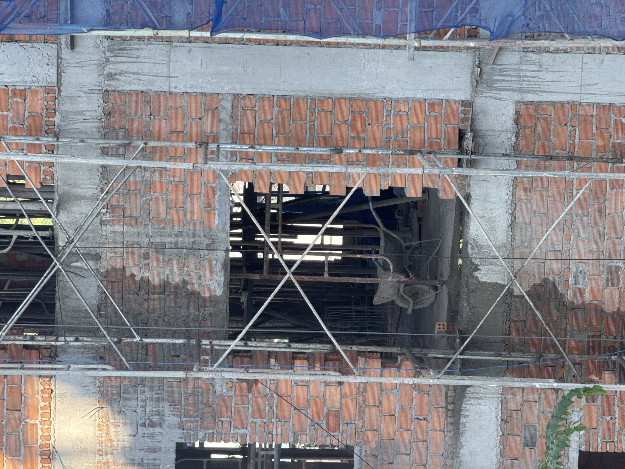 The image depicts the exterior of a multi-story building under construction in Bac Nha Trang Ward, Vietnam. The facade features a combination of red brick walls and concrete support columns. Scaffolding, constructed from metal poles and cross-braces, is erected around the building's exterior. A blue mesh safety net is visible at the top of the frame. The interior of the unfinished building is visible through the open spaces where windows or doorways will eventually be located, revealing additional scaffolding and construction materials.