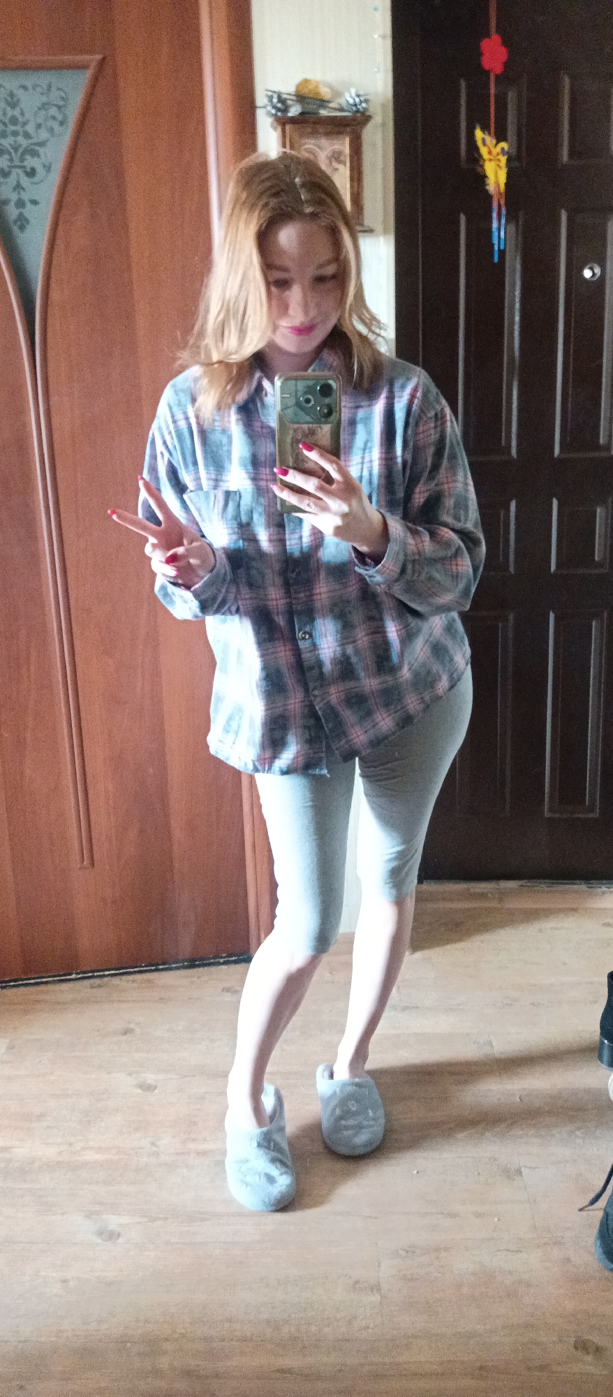 A full-body mirror selfie captures a person standing indoors. The individual, with light-colored hair, is wearing an oversized grey, pink, and light blue plaid long-sleeved shirt, light grey capri-length leggings, and light grey fluffy slip-on slippers. They are holding a smartphone with a dark case to take the picture, partially obscuring their face. Their left hand is raised, forming a peace sign (V-sign), and their fingernails are painted a vibrant red or pink. A slight smile is visible on their face. The background features a light wooden floor and two distinct wooden doors: one on the left with a decorative frosted glass panel, and a darker brown door on the right, possibly an entrance door, with a visible peephole. Above the dark door, a colorful red and yellow decoration, resembling a wind chime or mobile, hangs on the light-colored wall. A small decorative shelf with a framed picture and other items is also visible on the wall. The overall setting suggests a casual, indoor home environment, likely a hallway or entryway. There is no visible text.