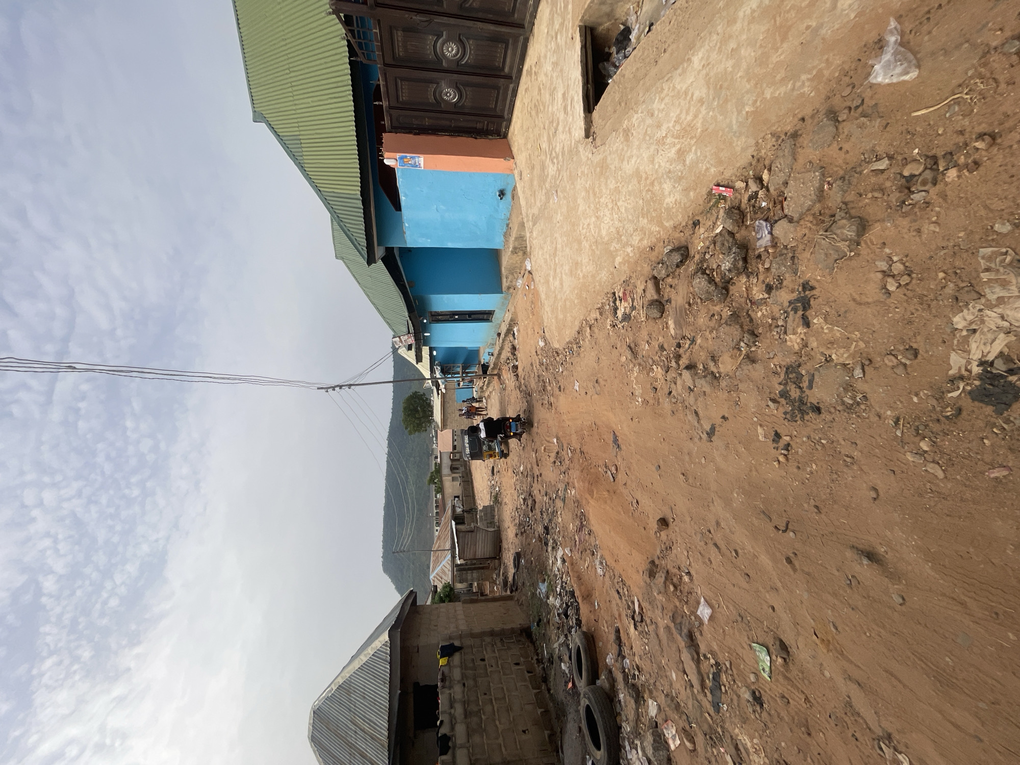 A daytime scene in Lokoja, Nigeria, depicts an unpaved, dusty road winding through a residential or commercial area, with a prominent mountain range in the background. The sky is largely overcast with scattered white clouds, suggesting diffused daylight.
The foreground and mid-ground are dominated by the dirt path, which is uneven and strewn with rocks, plastic debris, wrappers, and discarded tires. Buildings of varying construction line the path. On the left, structures feature corrugated metal roofs – one a light green, another a darker grey. A particularly visible building is painted in sections of bright blue and orange, featuring large, ornate dark brown double doors. A small, indistinct sign is visible on its blue wall.
Further along the path, utility poles and power lines are noticeable. In the middle distance, a small three-wheeled vehicle, possibly a commercial tricycle, is present on the path. Some figures, likely people, are visible near this vehicle, suggesting everyday activity. Text, "SHAH OF CLOTHES," is faintly discernible on the side of the three-wheeled vehicle.
The backdrop features a large, green mountain range under the hazy sky, adding a natural element to the bustling street scene. The overall impression is of a vibrant but unkempt community street, typical of many semi-urban environments in the region.