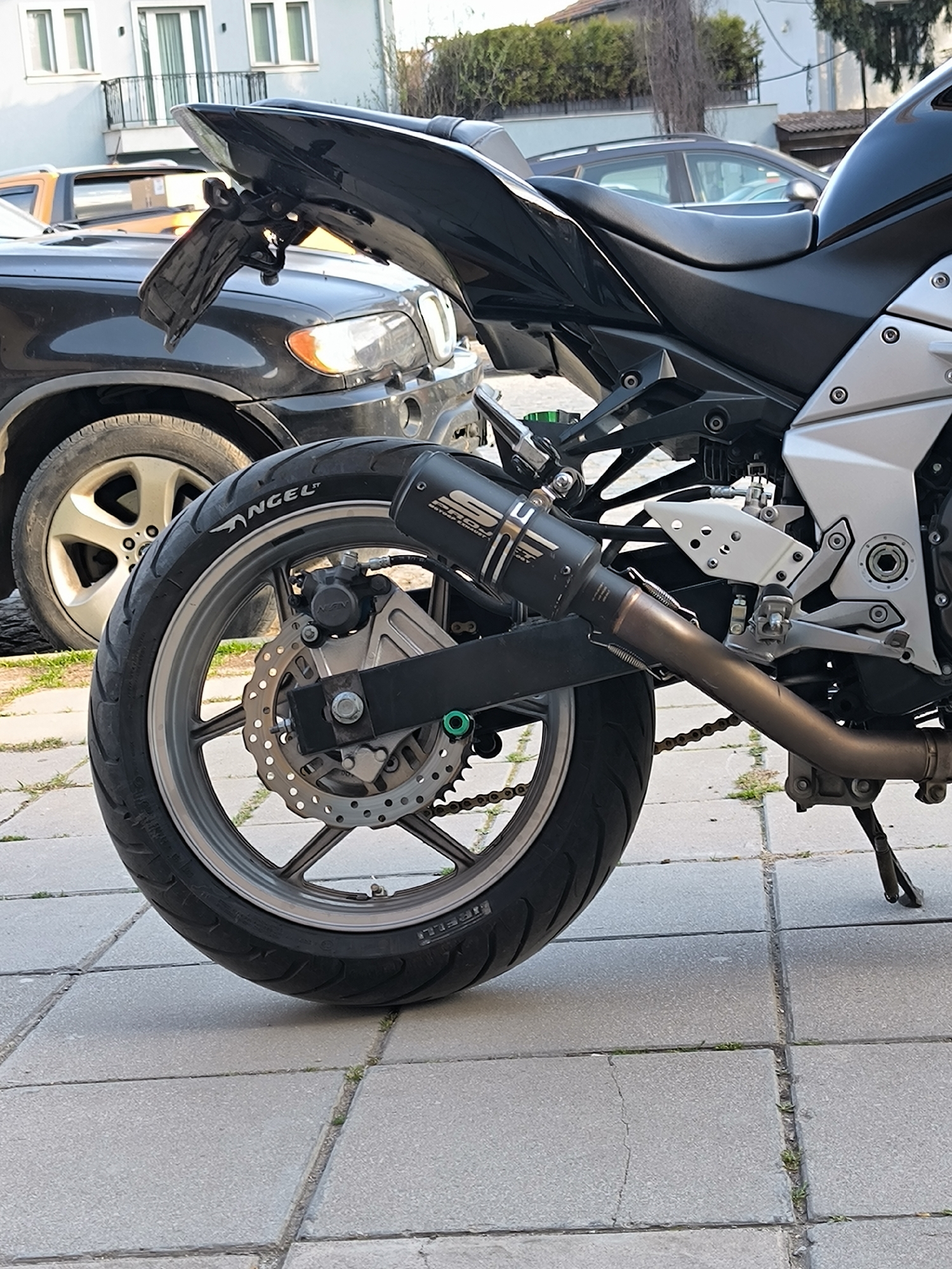 The image shows the rear section of a motorcycle, specifically the left side including the rear wheel, exhaust, and part of the frame. The motorcycle is parked on a paved surface composed of grey concrete blocks with small patches of grass growing in the joints. Behind the motorcycle, partially obscured, is a dark-colored BMW SUV. Further in the background are other vehicles and residential buildings. The location is suggested to be in Sofia, Bulgaria, based on the architectural style of the buildings and the type of paving. There are no people visible in the frame.