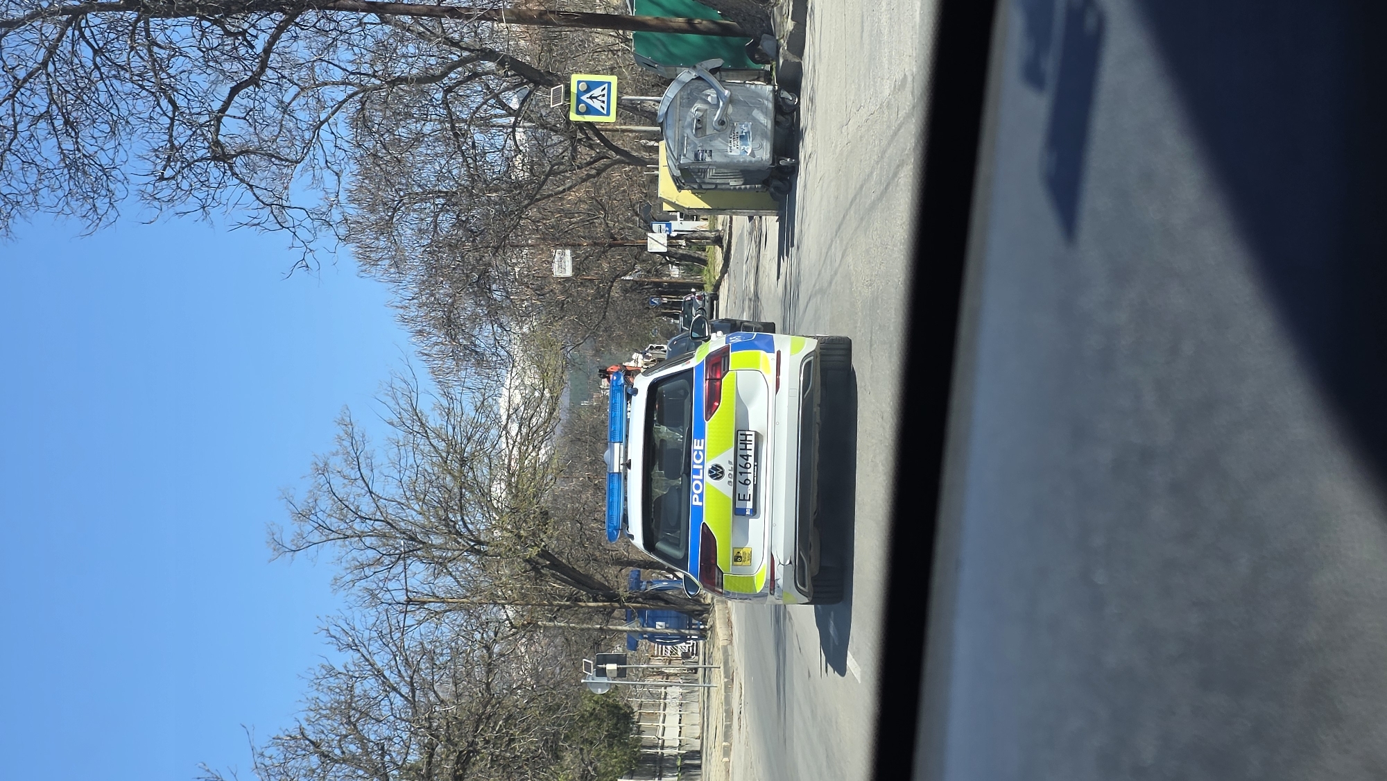 The image shows a white and blue Bulgarian police car with license plate number E-6164-HH on a road in Blagoevgrad, Bulgaria. The car is positioned in the middle of the lane, headed away from the camera. The background features trees without leaves and a bright blue sky. There is also a trash container and a pedestrian crossing sign visible on the right side of the road. The photograph appears to be taken from inside a vehicle.