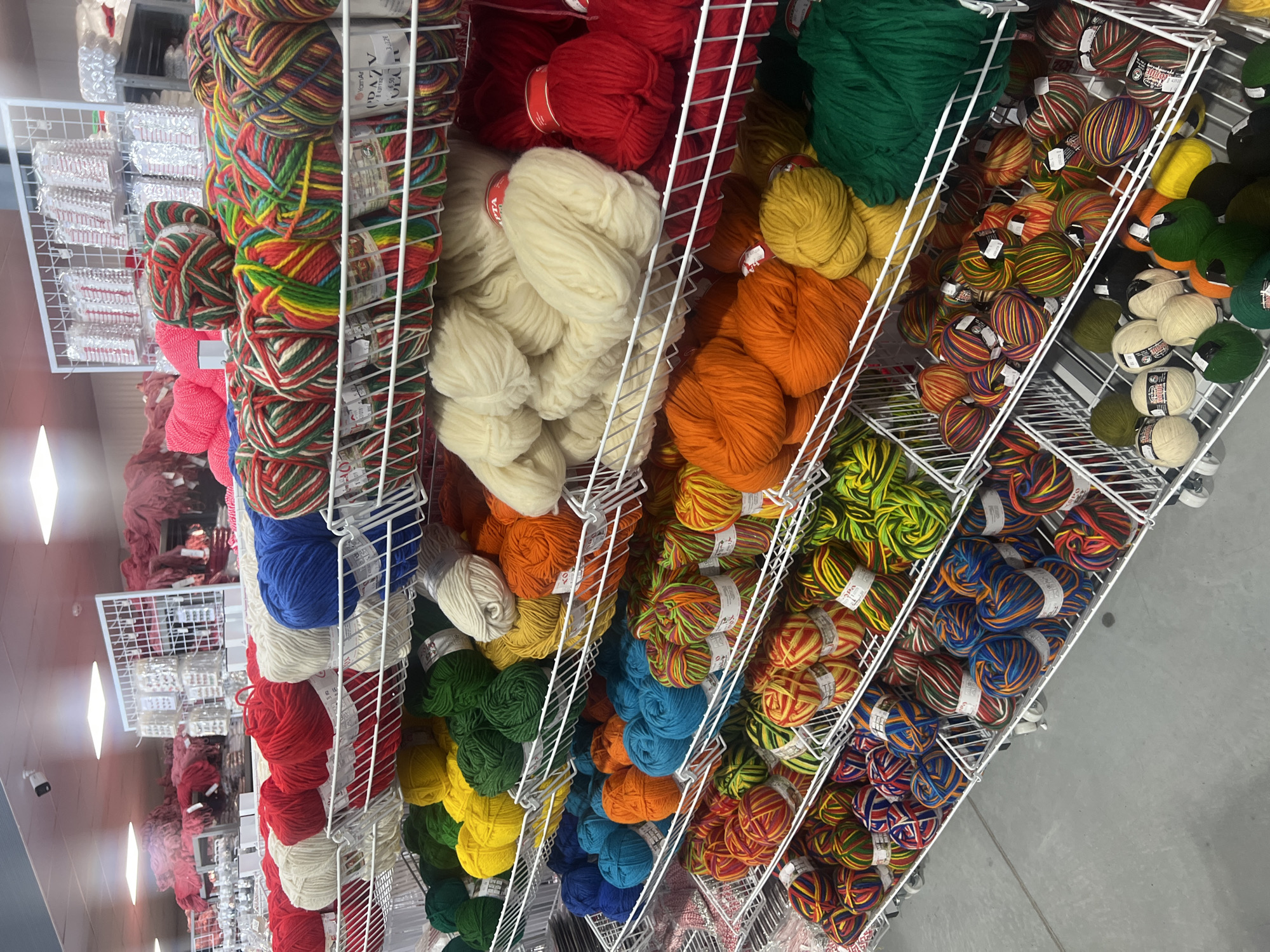 The image shows a retail display rack filled with balls of yarn. The rack is made of white wire and has multiple shelves. The yarn is arranged by color. The colors present are red, white, green, orange, yellow, blue and several multicolored varieties. The location is in the city of Burgas, Bulgaria.