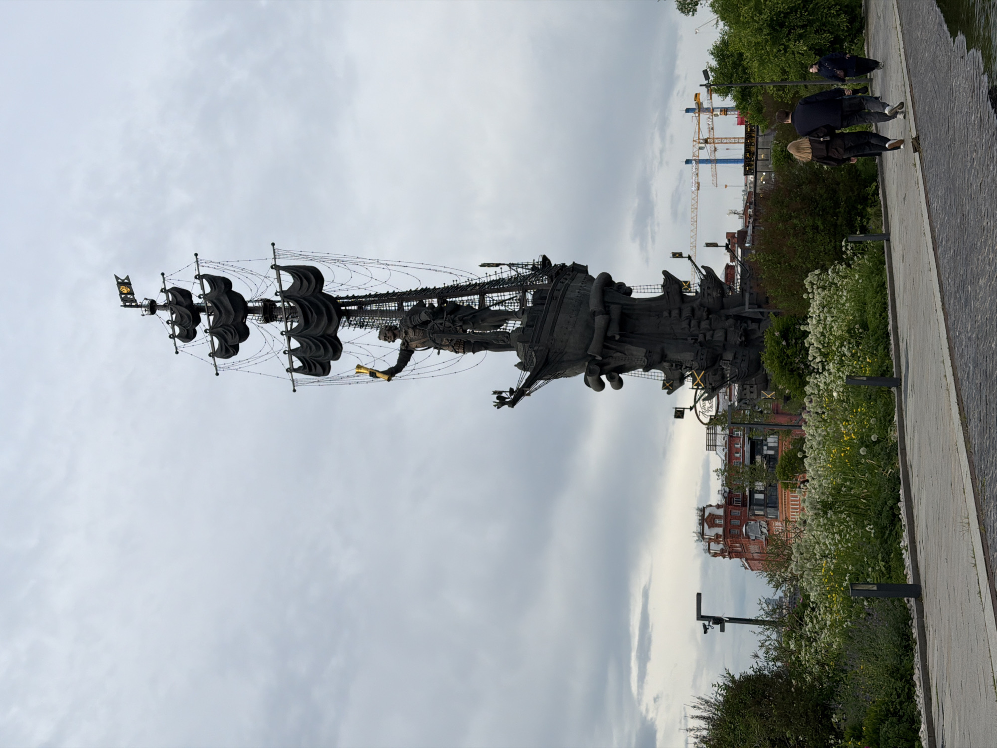 The image shows the Peter the Great Statue in Moscow, Russia. The statue depicts Peter the Great standing on a ship with sails. The sky is cloudy. People are walking on a path next to the statue. Construction cranes are visible in the background. There are buildings and greenery in the foreground.