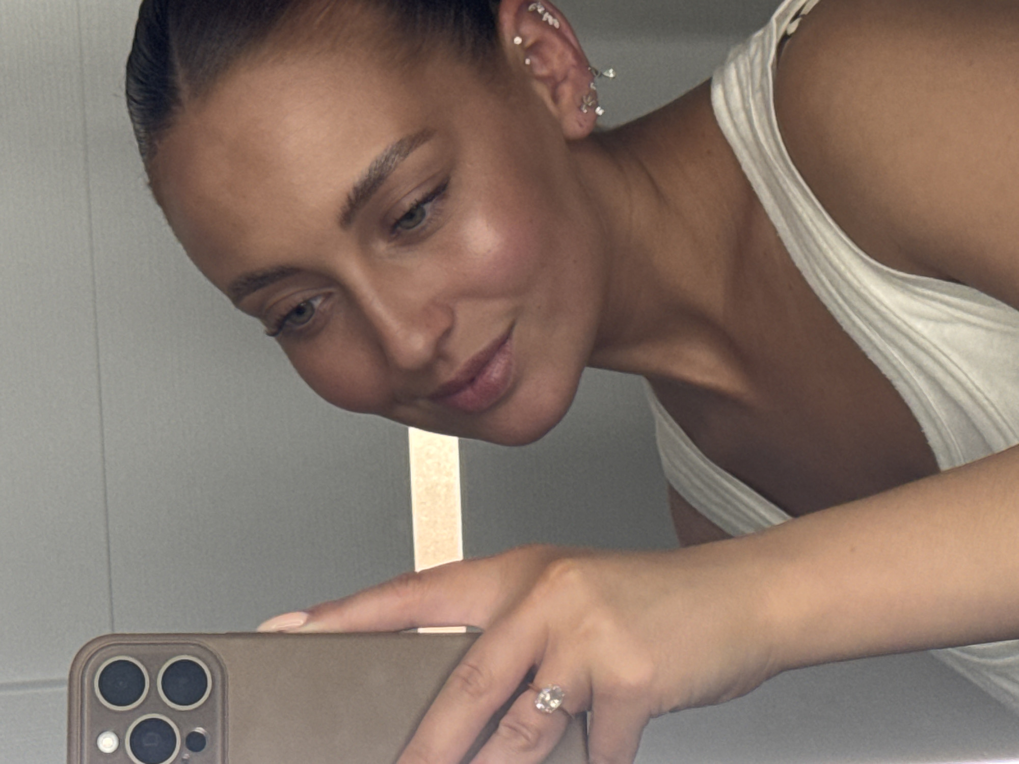 A woman is taking a selfie in a mirror. She is holding a smartphone in her left hand. She is wearing a white tank top. She has on a ring with a large stone on her left ring finger and multiple earrings in her right ear. She has her hair pulled back. The background appears to be a bathroom with grey tiled walls. The image is a close-up and the woman is the only person visible.