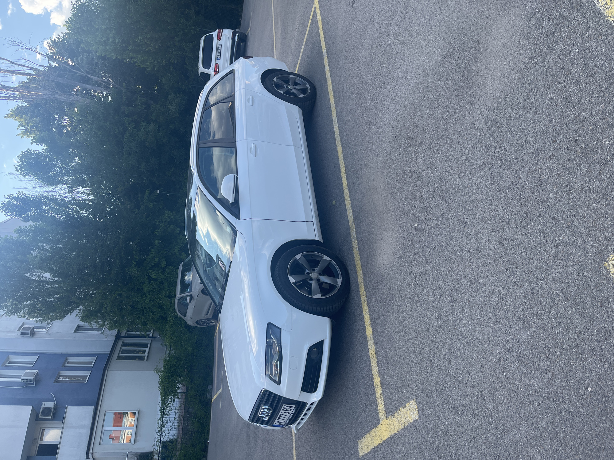 This media file depicts a white Audi A4 Avant parked in an outdoor parking lot, viewed from a slightly elevated side angle. The car is the primary subject, with its front and side prominently visible. The license plate reads "PK 773 BX". The parking lot is marked with yellow lines, and the asphalt surface is visible. In the background, there is a dense green tree line and a residential building with blue and white facade. The sky is partly cloudy, suggesting it is daytime. The overall setting appears to be urban, likely a residential area. There are no people visible in the image. The scene is static, with the car parked and no activity taking place. The lighting and shadows indicate it is likely late afternoon or early evening. The location context is identified as Sofia, Bulgaria.