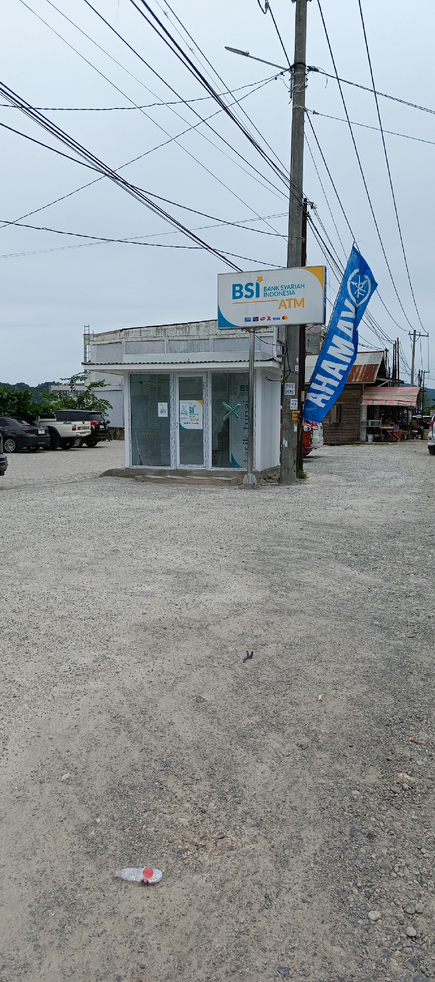The image captures an outdoor scene in Lampeuneurut Blang, Indonesia, dominated by an ATM kiosk for Bank Syariah Indonesia (BSI). The kiosk is a small, modern structure with large glass windows and a white roof, situated on a gravel lot. Above the kiosk, a prominent BSI sign advertises "BANK SYARIAH INDONESIA ATM" and displays various payment card logos. A blue Yamaha flag flutters on a pole next to the sign.
In the background, several vehicles are parked, including a black sedan, a white SUV, and another dark-colored vehicle. Beyond the parked cars, there are some trees and buildings, including a rustic structure with a corrugated metal roof. Overhead, a dense network of electrical wires crisscrosses the sky, attached to a utility pole that supports the BSI sign and a street lamp. The ground is covered in a coarse gravel surface. In the foreground, a discarded plastic water bottle with a red cap lies on the ground, and a small, unidentifiable dark object is also visible. The sky is overcast, suggesting a cloudy day.