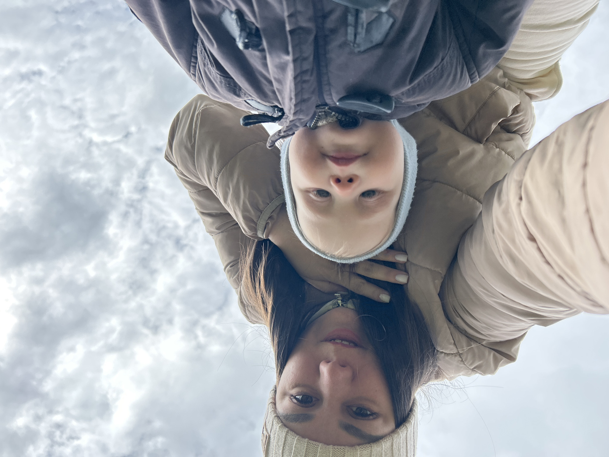 From a low-angle perspective against a cloudy sky, the image shows a woman and a child, both in winter attire. The woman is wearing a light-colored beanie and a beige puffer jacket. The child, positioned above the woman, wears a gray winter hat and a brown puffer jacket. The subjects appear to be taking a selfie. The specific location within the city of Chaikovsky, Russia cannot be identified from the visual information provided.