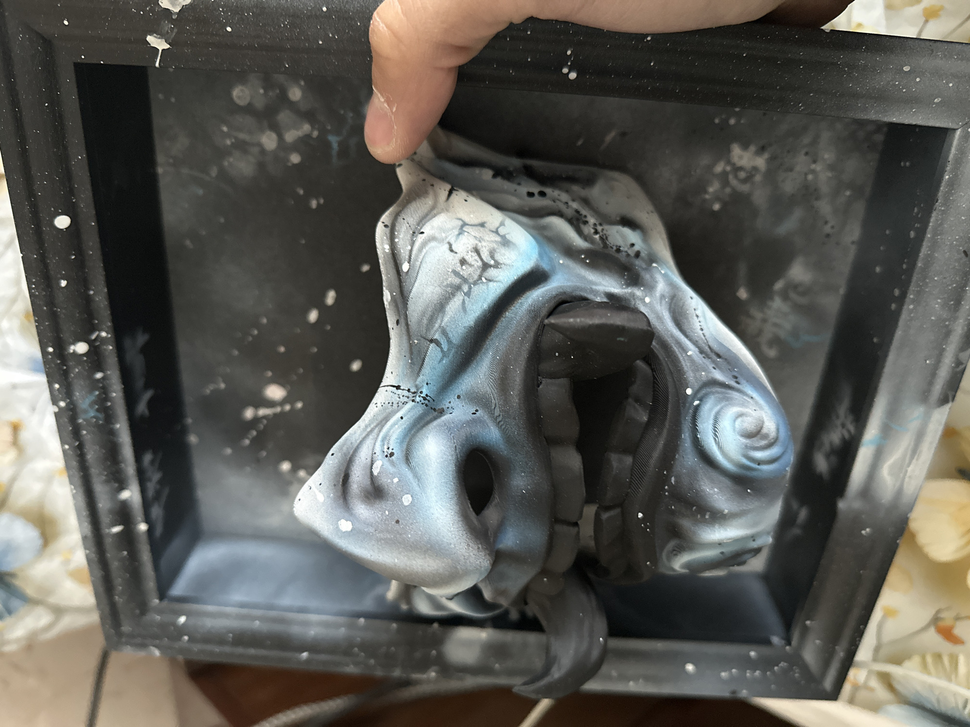 A hand holds a sculpted, painted mask within a dark-colored, painted shadow box. The mask is predominantly white and blue with black accents, featuring swirling patterns and textures. The background inside the box is a mottled gray and black with white splatter-like markings. The box itself has a speckled, painted finish, possibly suggesting a celestial theme. The scene appears indoors, with a glimpse of floral patterned fabric in the background. There is no discernible connection to Burgas, Bulgaria in the image.