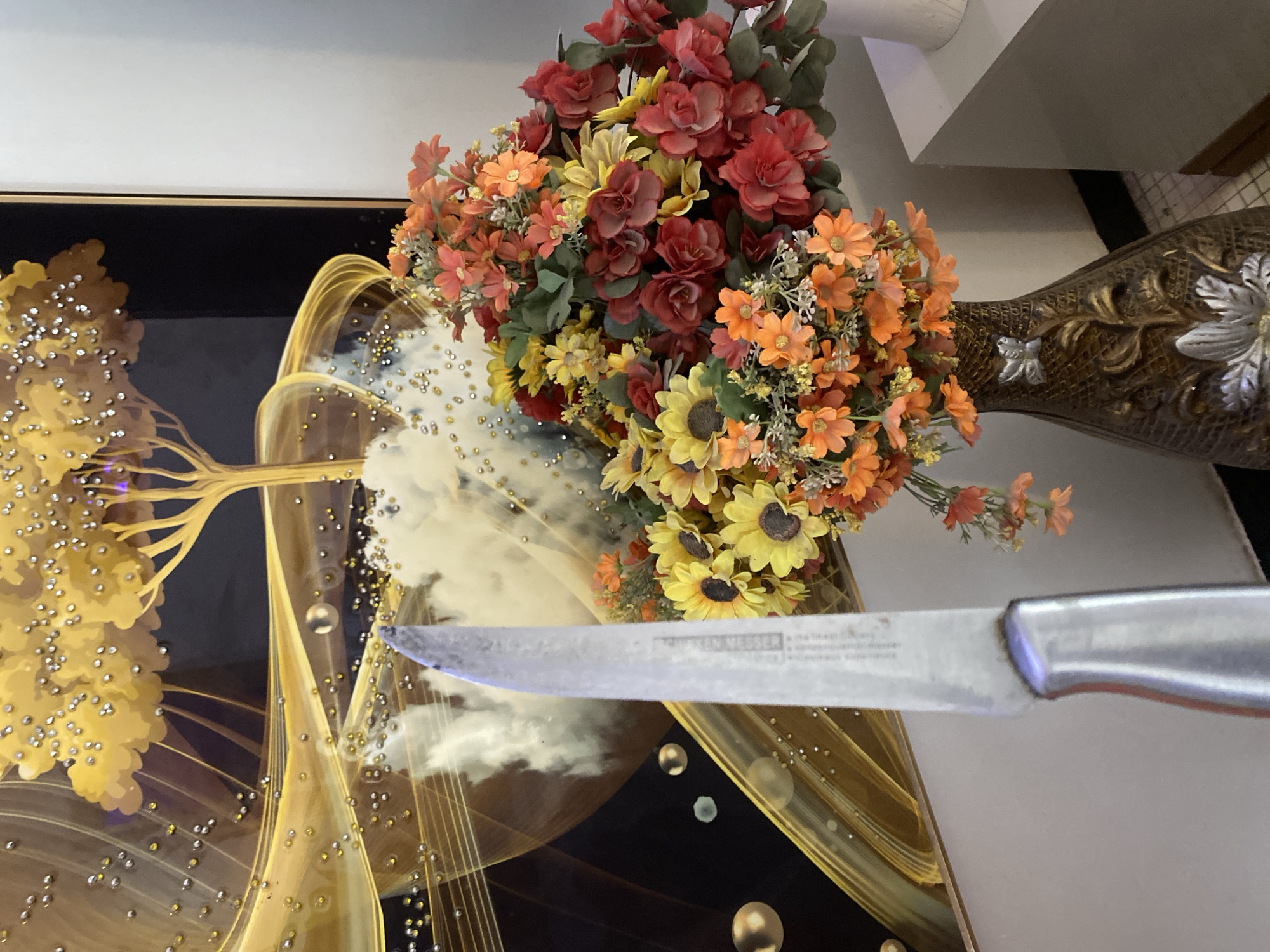 A close-up shot captures a vibrant artificial flower arrangement in shades of red, orange, and yellow, spilling out of a decorative vase. In the foreground, the blade of a silver knife is prominently displayed, its edge angled towards the viewer. The knife appears to have some text etched onto its surface, though it is partially obscured. Behind the flowers and knife, a piece of art or framed picture is visible. This artwork features abstract golden swirls and patterns, interspersed with what look like clusters of small, reflective beads, resembling dew drops or tiny jewels. The background is a plain, light-colored wall, and a hint of a tiled surface is visible at the bottom right. The lighting suggests an indoor setting, likely during the daytime. The overall impression is a still life composition, blending natural elements with decorative objects.