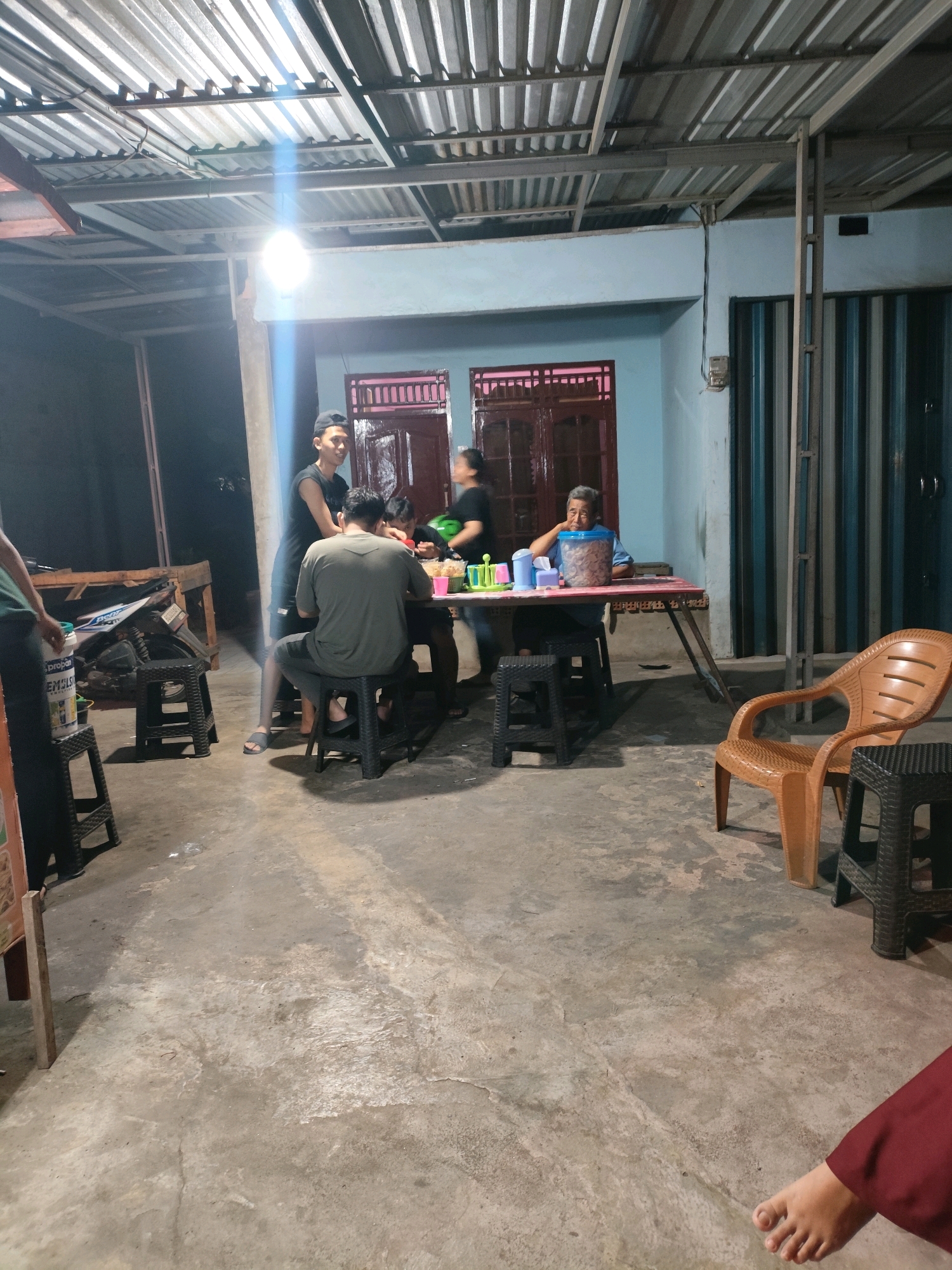 The image depicts an outdoor gathering at dusk or night, possibly a casual eatery or home in Palembang, Indonesia. A corrugated metal roof with exposed beams and lighting covers the scene. In the center, a group of people are seated around a table. One man, wearing a grey t-shirt and shorts, is seated facing away from the camera, looking down at the table. Another man, wearing a black t-shirt and cap, stands behind him, seemingly interacting with the group. To the right, an older man in a blue shirt leans on the table, looking thoughtful. A woman in a black t-shirt is visible in the background, tending to the table. The table is laden with food items in containers, including a large jar of what appears to be snacks. In the foreground, black plastic stools are arranged around the table, and a brown plastic chair is visible to the right. The ground is a rough concrete surface. To the left, a motorcycle is parked next to some wooden structures and a large white cylindrical container. The overall impression is a relaxed, communal meal or gathering.
