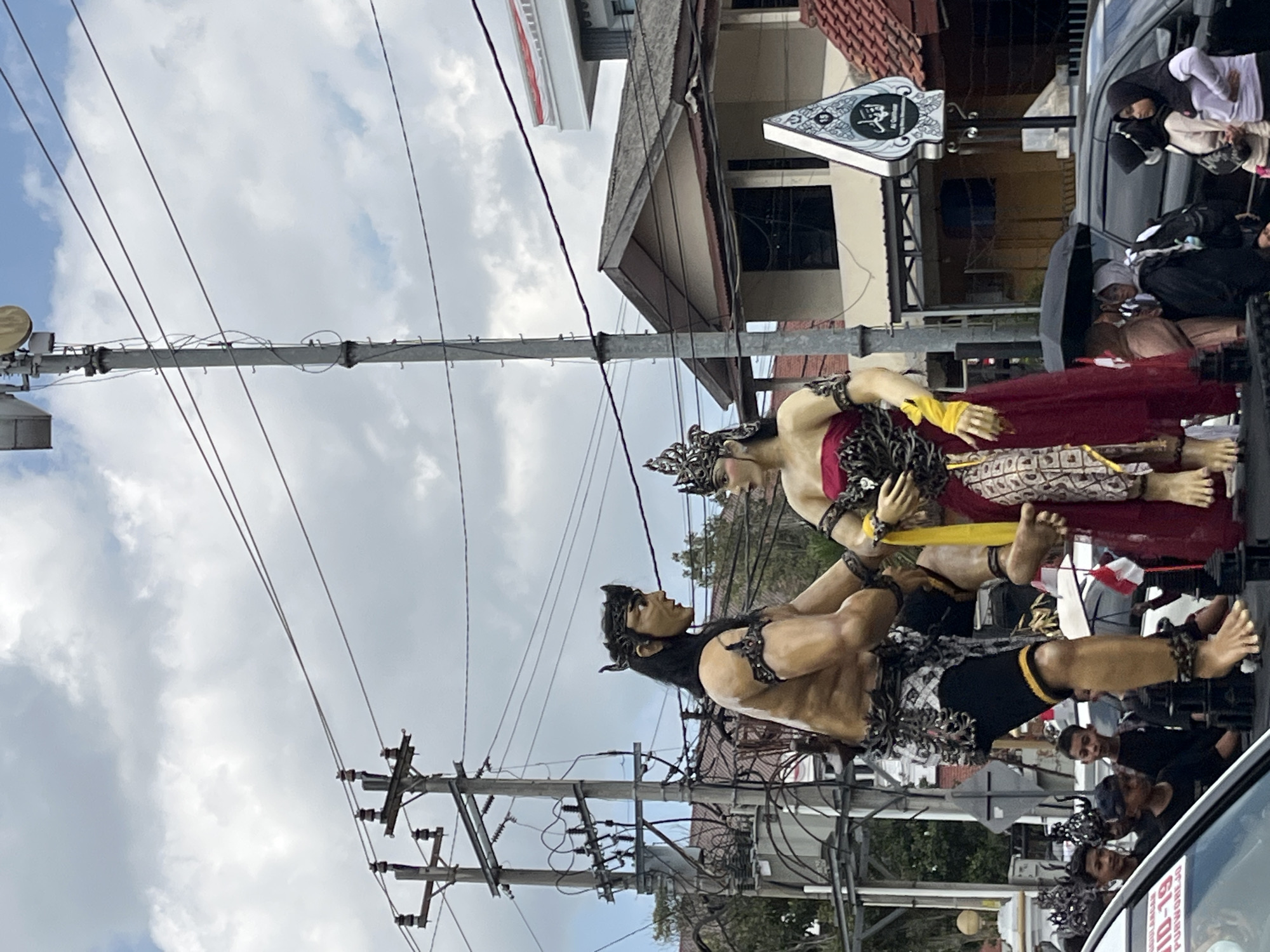 In Unknown, Unknown, a cloudy sky looms over an outdoor street scene. Dominating the foreground are two large, elaborate statues of figures in traditional attire. The male figure, positioned on the left, is muscular and wears a dark headdress, a beaded chest ornament, and a patterned loincloth with black shorts. He holds a female figure, who is seated on his lap. The female figure is adorned with a crown, a strapless red dress with beaded detailing, and a yellow sash. Her attire includes a patterned skirt.
Behind the statues, a utility pole with numerous wires and insulators stands prominently, suggesting an urban or semi-urban environment. In the middle ground, a multi-story building with a slanted roof and dark-paned windows is visible. Further back, another building with a white facade and a red-and-white striped banner is partially seen.
A procession or gathering is implied by the presence of several people in the lower part of the image. Some are looking towards the statues, while others appear to be part of the event. Several cars are also present, including one in the foreground with part of its hood and windshield visible, displaying text that includes "PURWOREJO". Umbrellas, one black and one red and white, are being used by some of the attendees, indicating potential light rain or strong sun. The overall atmosphere suggests a cultural event or celebration taking place on a street.