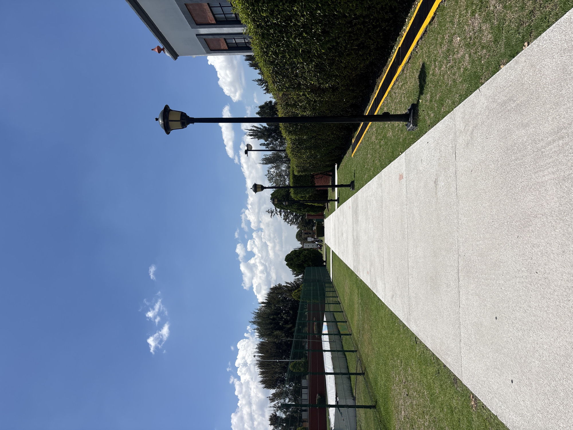 The image shows a paved walkway in San Andrés Cholula, Mexico, lined with grass and bushes on one side, and a green fence with a tree line behind it on the other. Black lamp posts are spaced along the walkway. A building is partially visible at the top, and the sky is blue with scattered clouds.