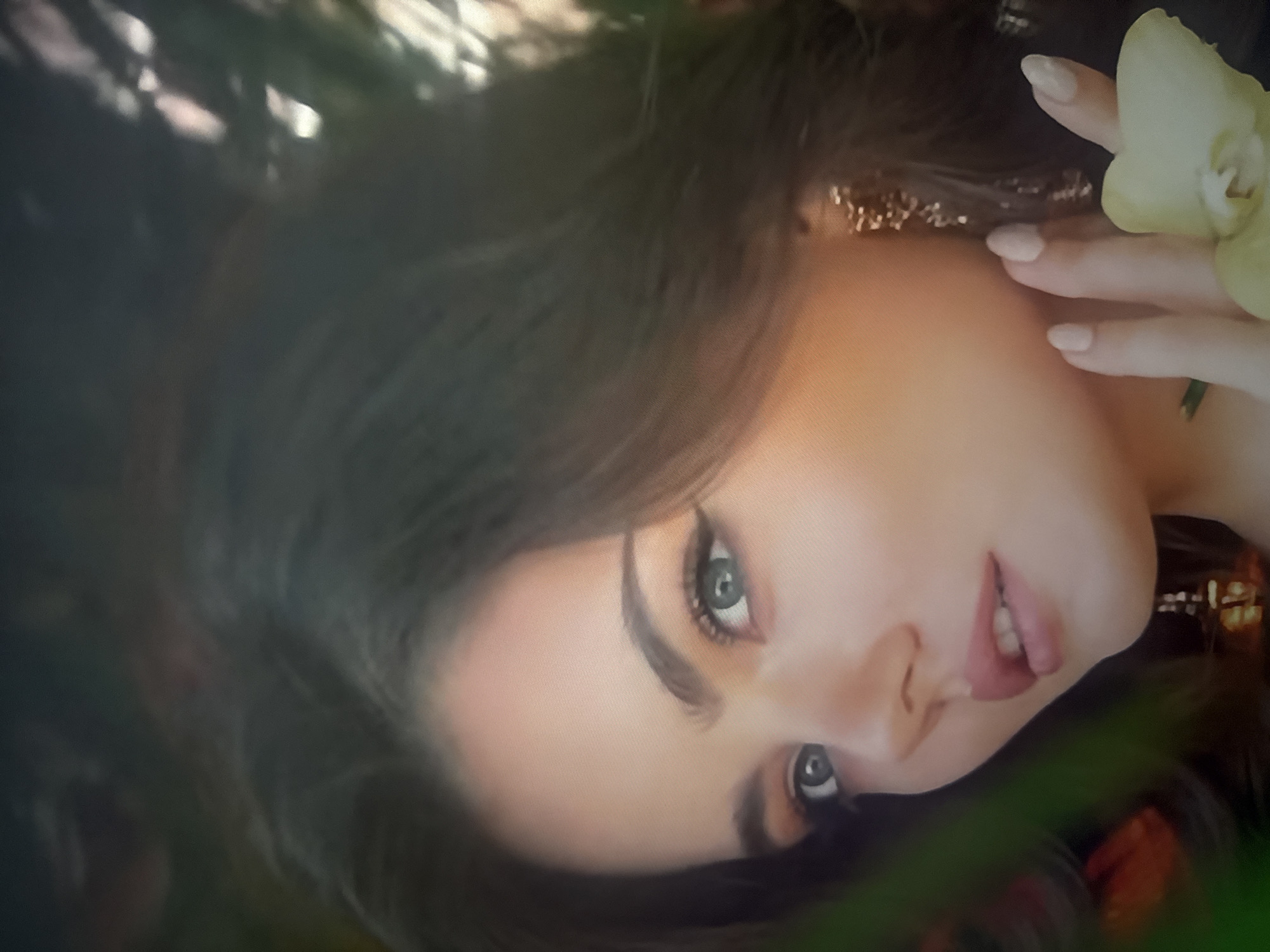 The image shows a close-up of a young woman's face, positioned in the foreground. She has dark hair, fair skin, and light blue eyes. Her left eye is looking directly at the camera, while her right eye is partially obscured. She is wearing eye makeup and lipstick. To the right of her face, her hand is visible, with manicured fingernails, holding a light yellow flower. She is wearing ornate, dangling earrings. The background is dark and appears to be foliage, with hints of orange and green. The image is taken from a slightly low angle. There is no identifiable location within the city of Herzliya, Israel in this image.