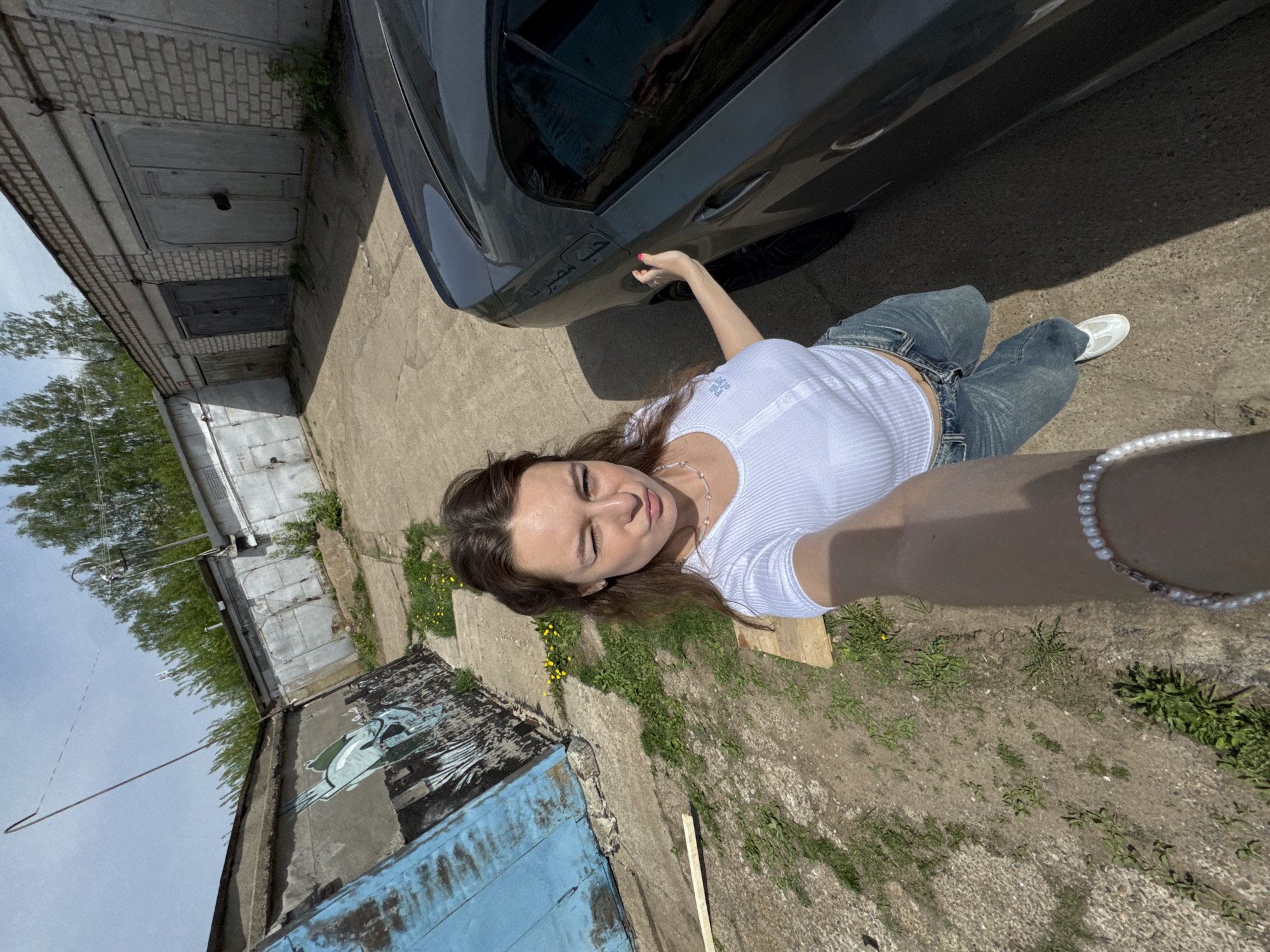 The image depicts a young woman taking a selfie in what appears to be an alley or yard near a row of garages. She is wearing a white top and blue jeans, with a pearl bracelet visible on her extended arm. A dark-colored car is parked beside her. The ground is a mix of concrete, dirt, and grass. One of the garages has blue paint and a mural on its door. The location is Chaikovsky, Russia.